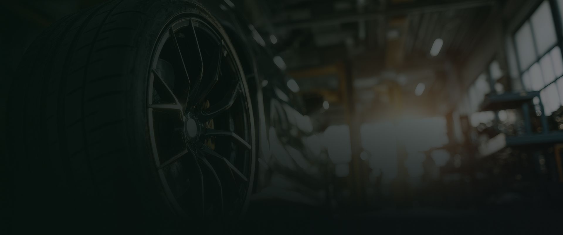 Dimly lit workshop with a large wheel or machine part in the foreground and a bright light in the background | OG Autoworks