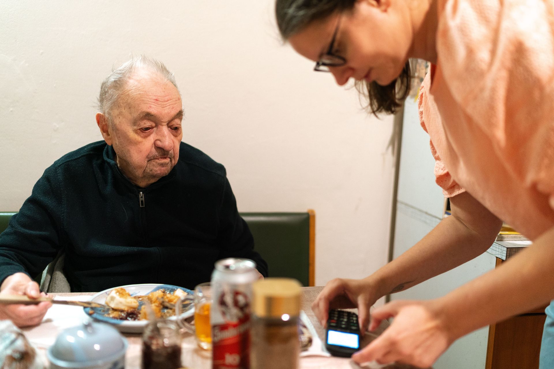 Person serving food to an older man at a dining table while taking a photo with a phone