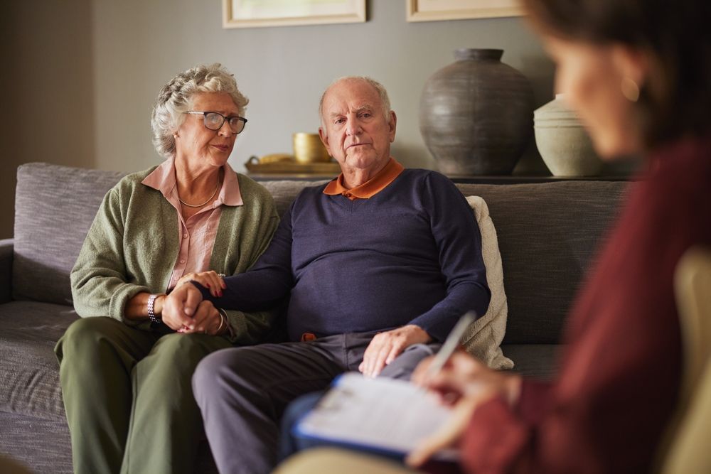 Older couple sitting on a couch, listening to a counselor in a living room.
