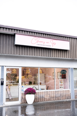 Small storefront with large windows, a white sign, and a white planter of purple flowers by the entrance.
