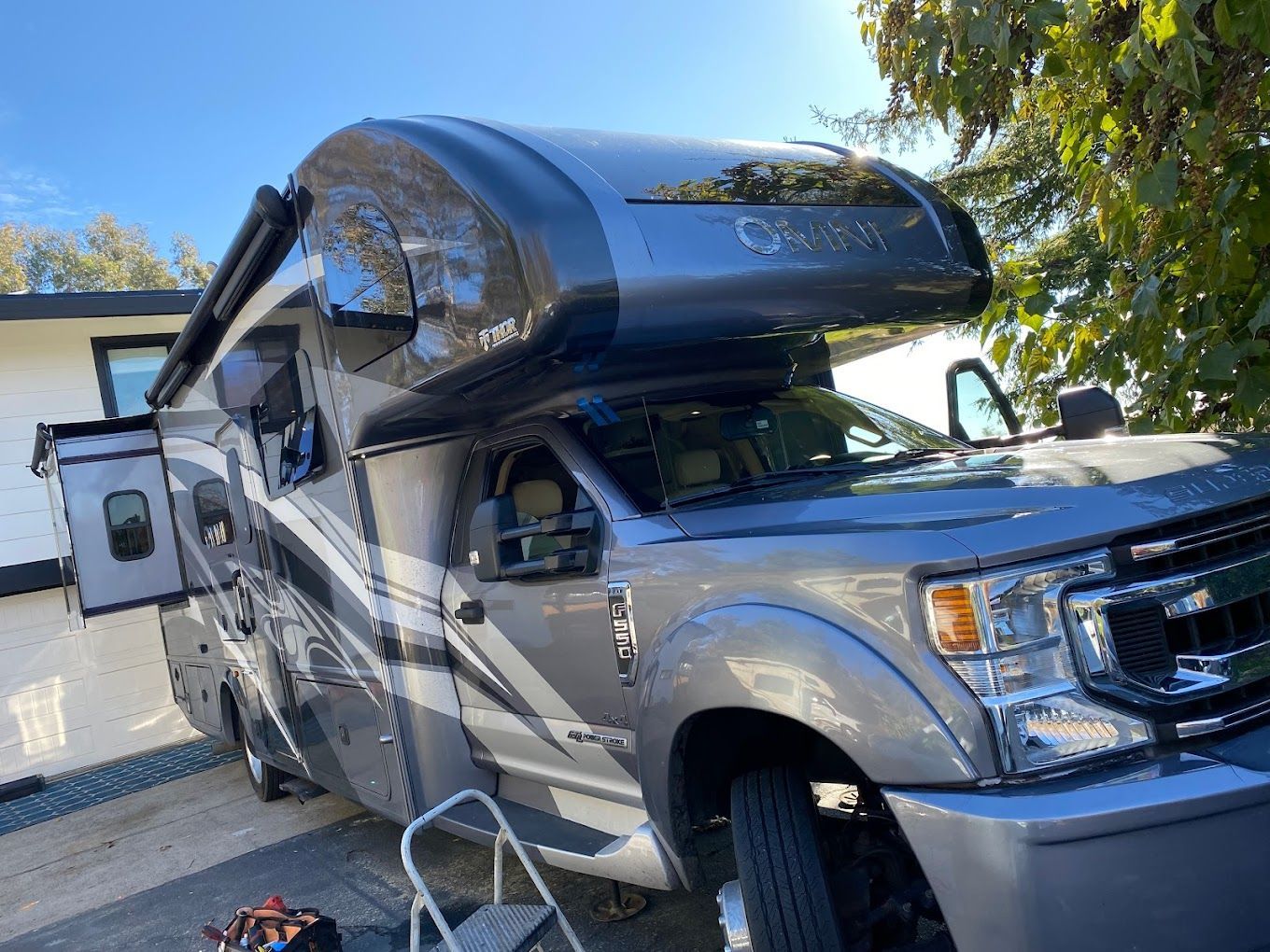 Silver RV parked in a driveway beside a white garage, with a raised black roof and front steps.