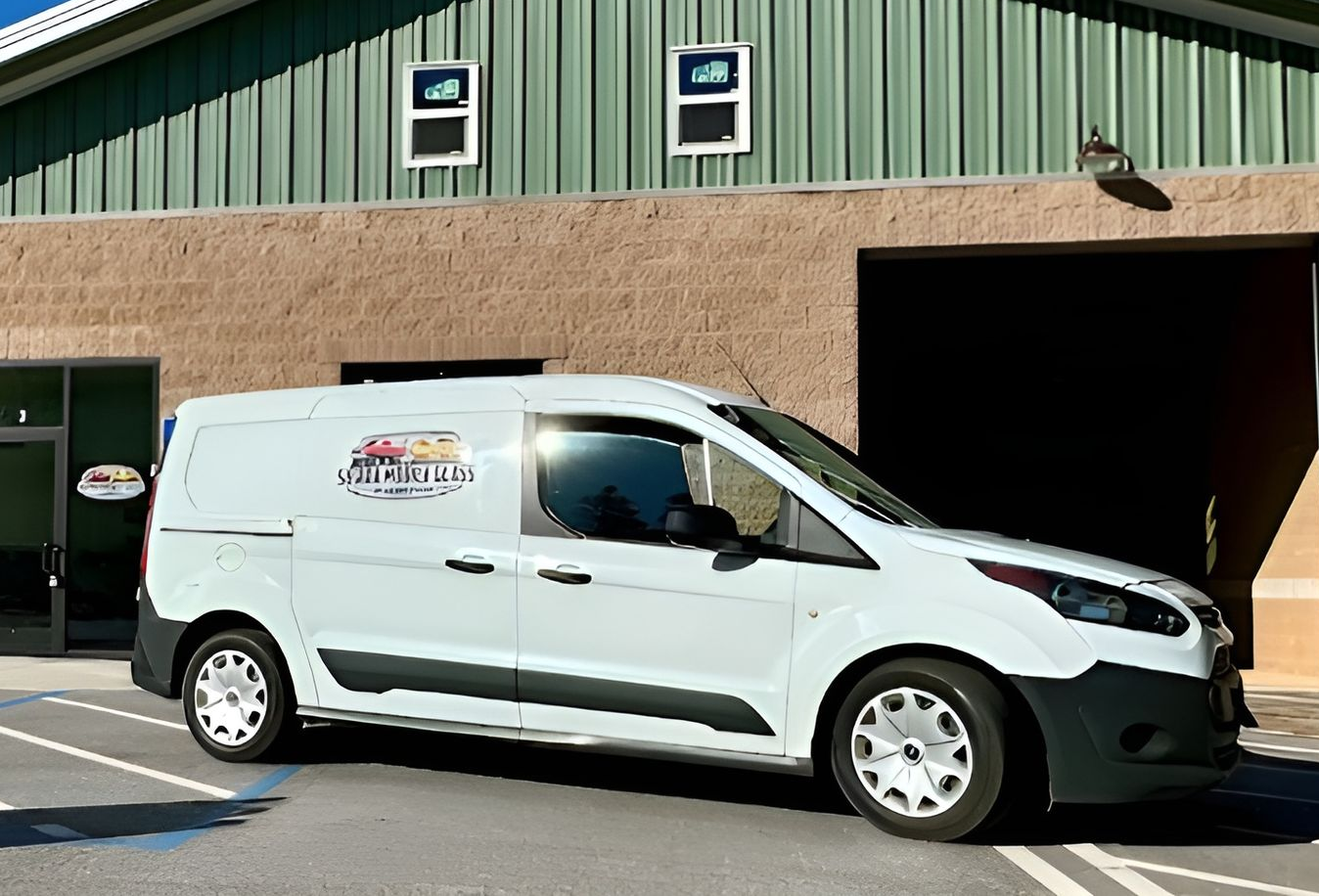 White service van parked outside a green and tan industrial building with open garage bays.