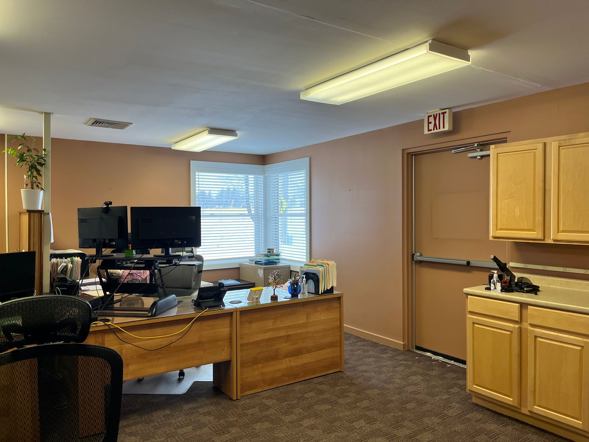 A well-lit office with a wooden desk, computer monitors, a rolling chair, beige walls, and wood cabinets.