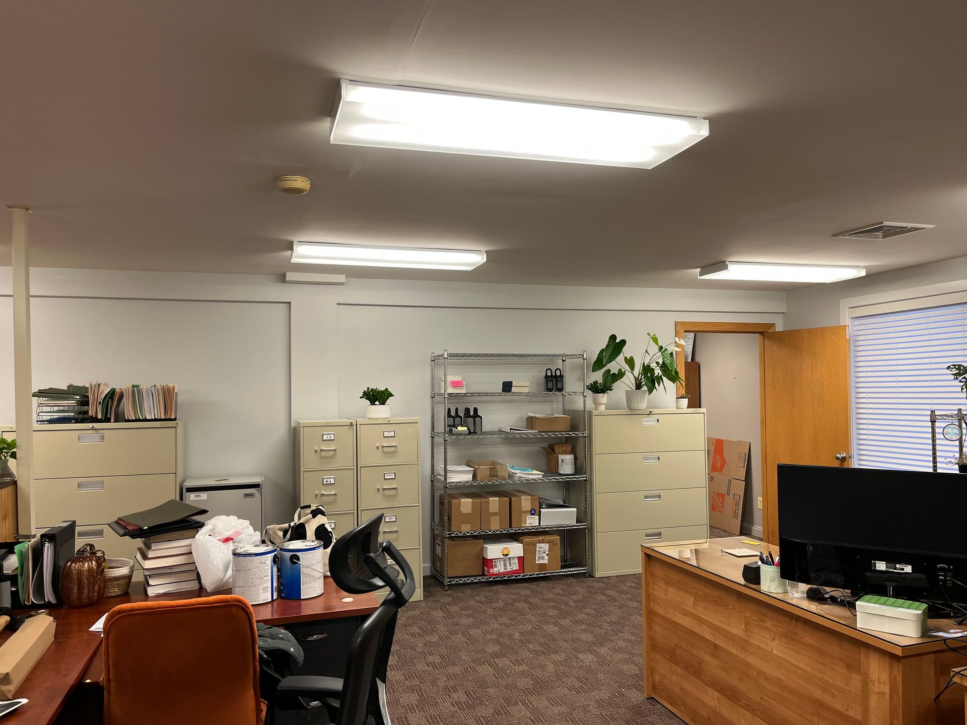 An office space featuring filing cabinets, a metal shelving unit with supplies, desks, and overhead fluorescent lighting.