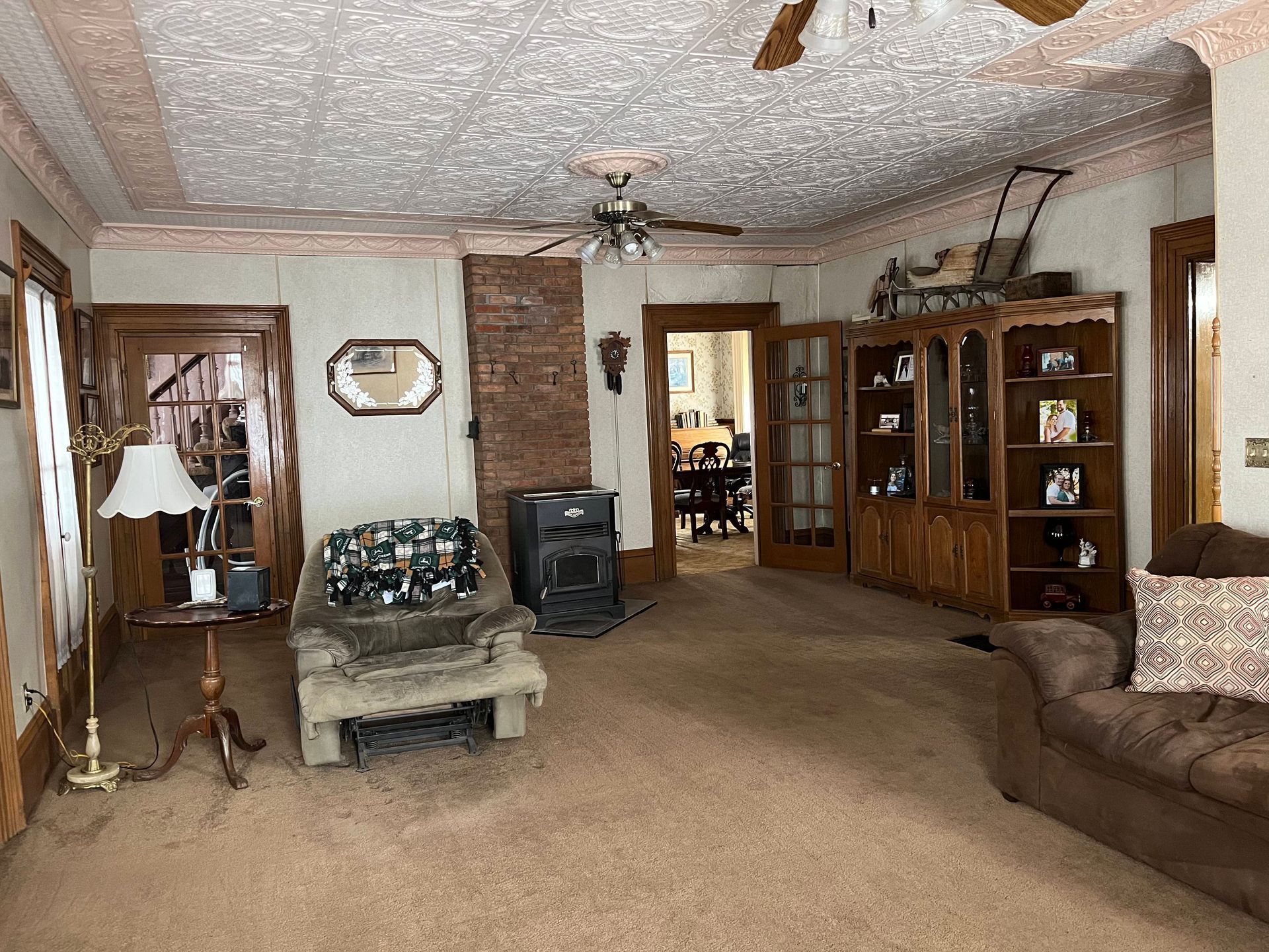 A living room featuring a patterned ceiling, a brick fireplace with a wood stove, tan carpeting, and wooden shelving.