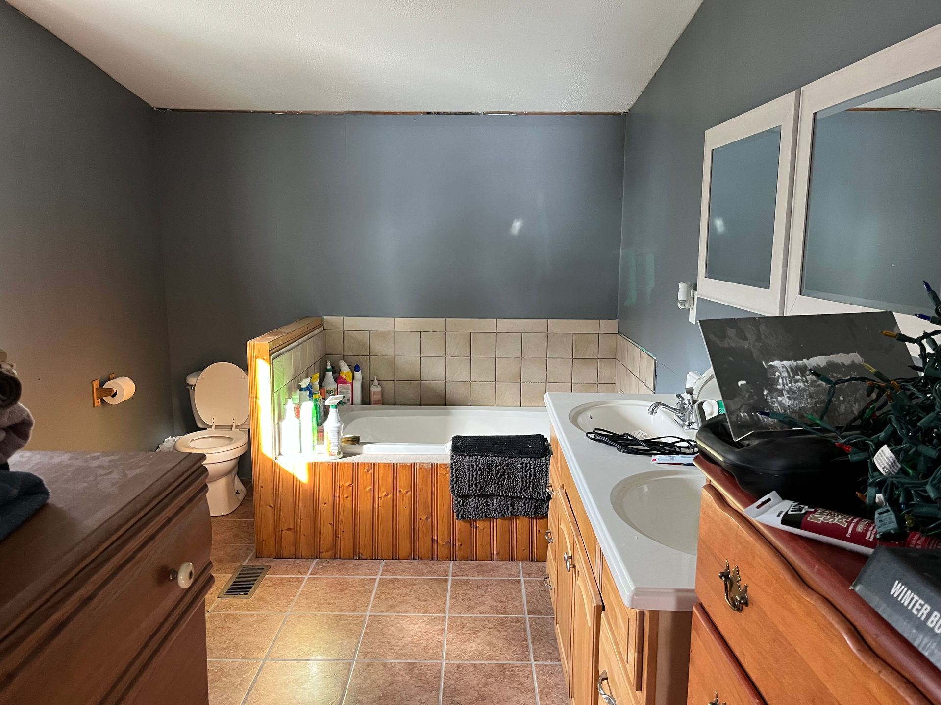A bathroom with a dual-sink wooden vanity, tile floor, dark gray walls, a toilet, and a tiled soaking tub with wood trim.