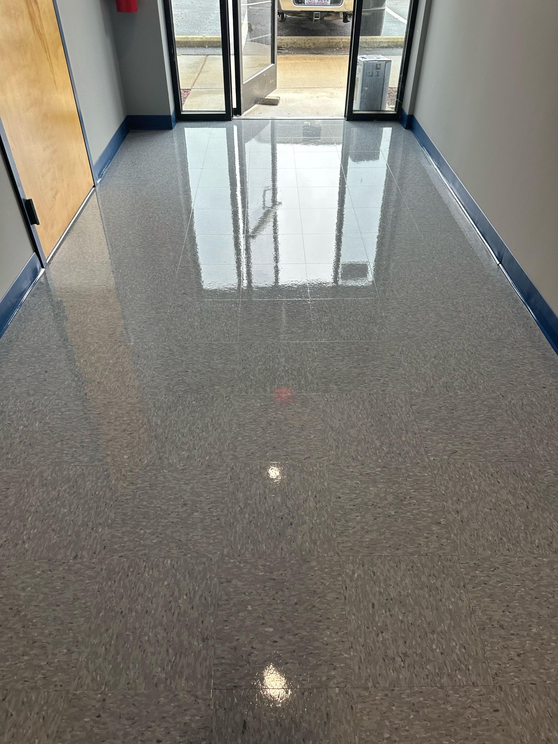 Shiny, speckled gray floor in a hallway with glass doors at the end. Blue tape lines the edges.