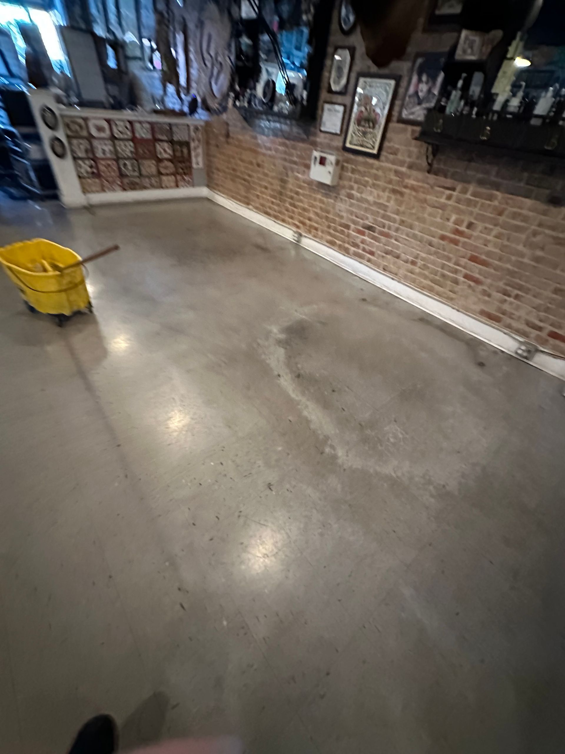 A yellow bucket and a mop sit on a polished concrete floor. Brick wall in the background.