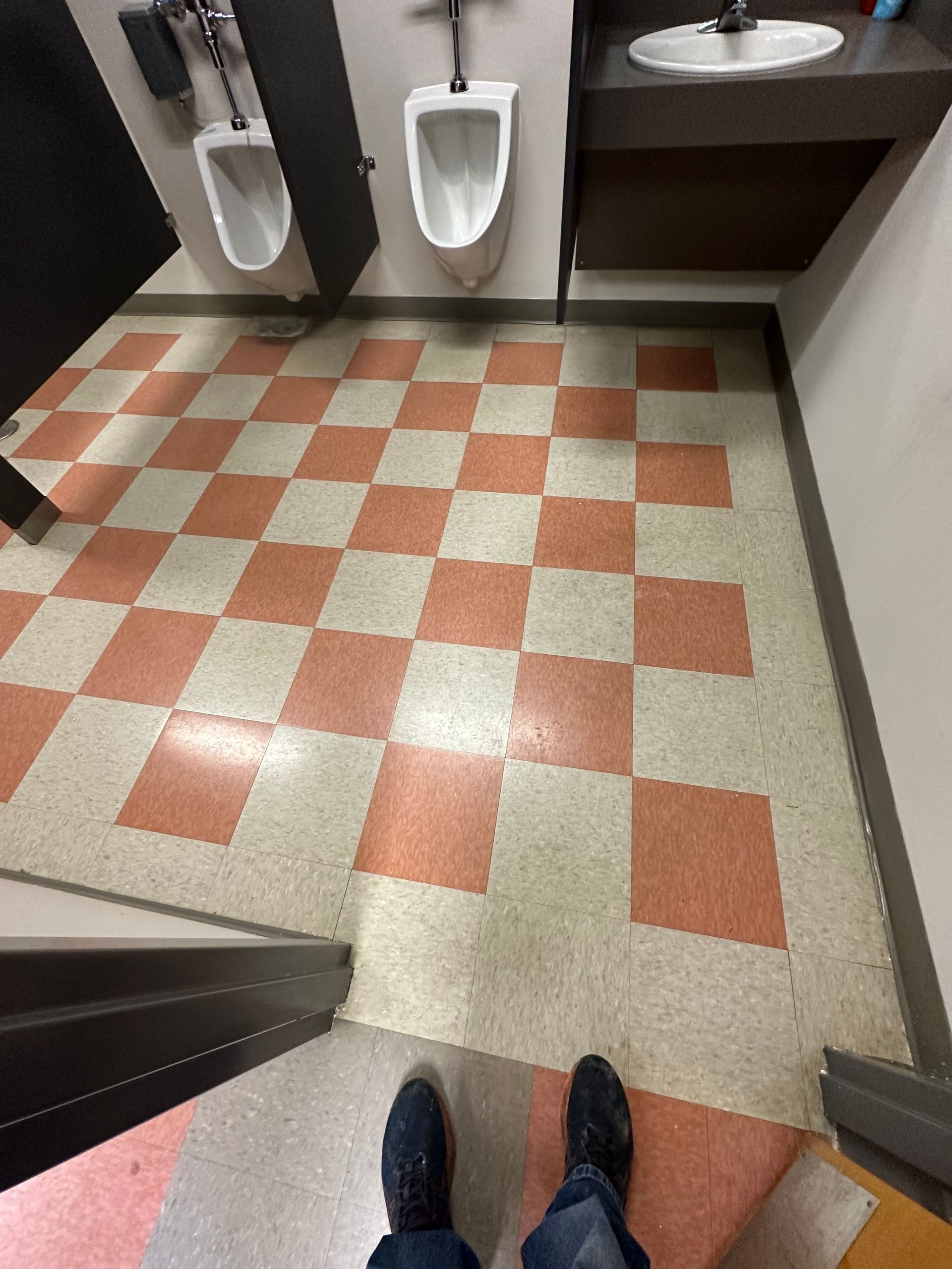 A person's feet in blue jeans and black shoes in a public restroom with red and white checkered floors, urinals, and a sink.
