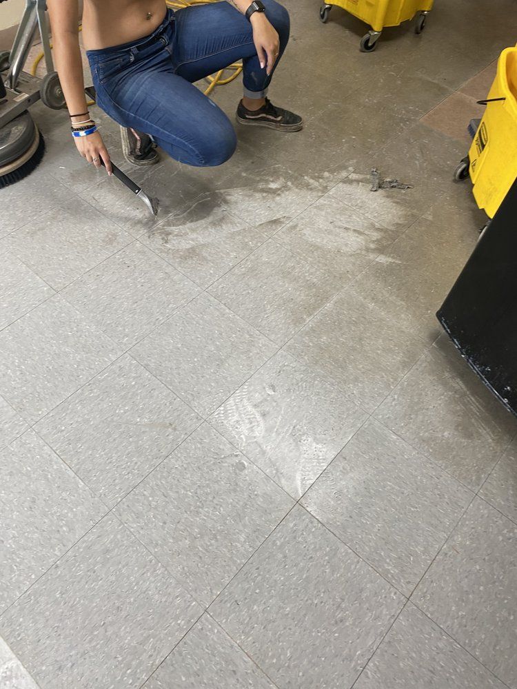 Person kneeling, scraping something off gray floor. In a school setting.