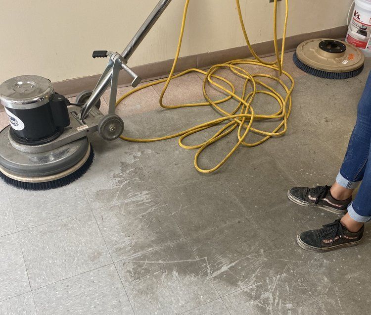 Floor cleaning machine in use, person operating it. Yellow cord, light gray floor, beige wall.