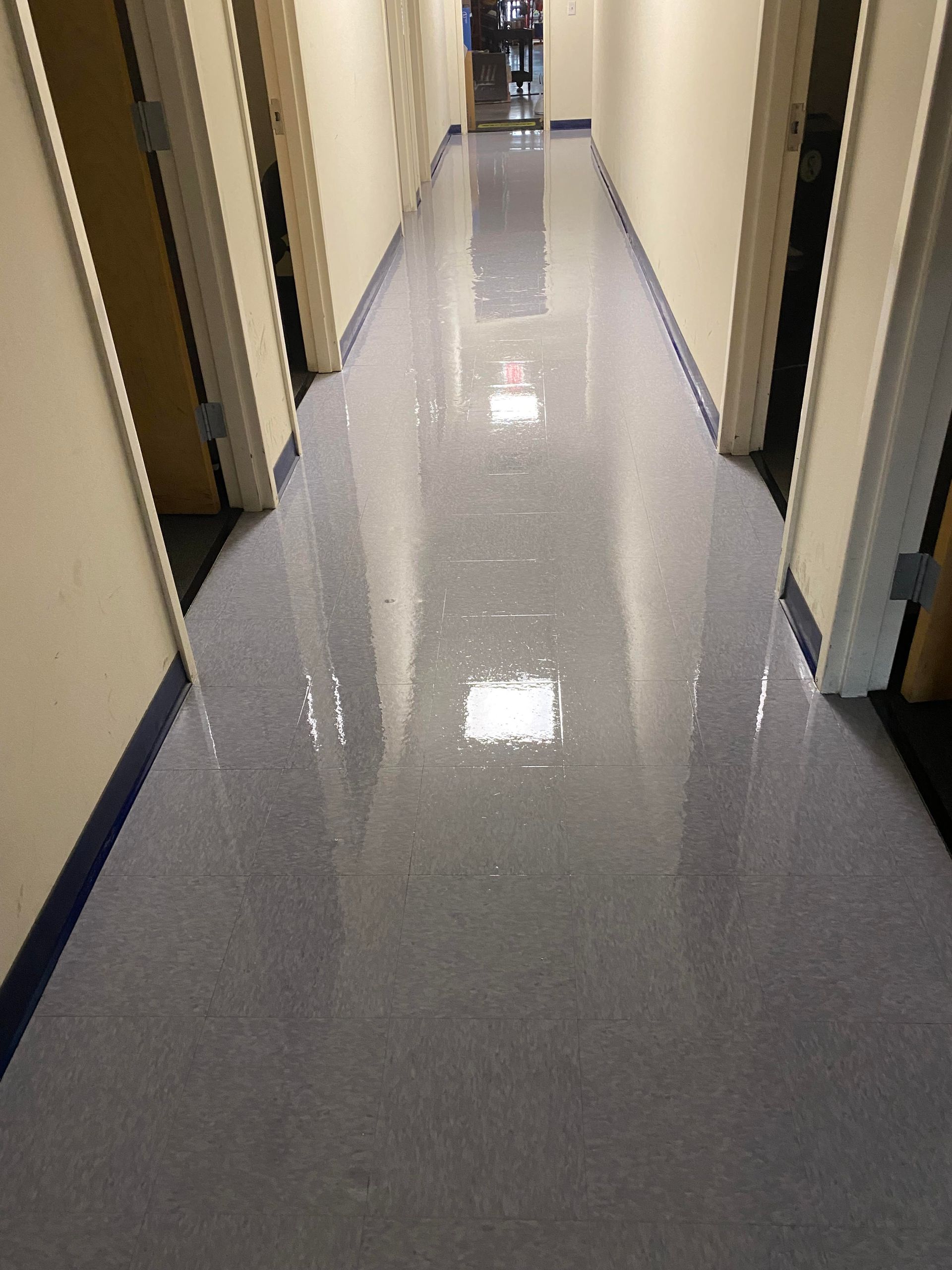 Long hallway with shiny, light-gray flooring. Doors line both sides, and a dark blue border runs along the base of the walls.