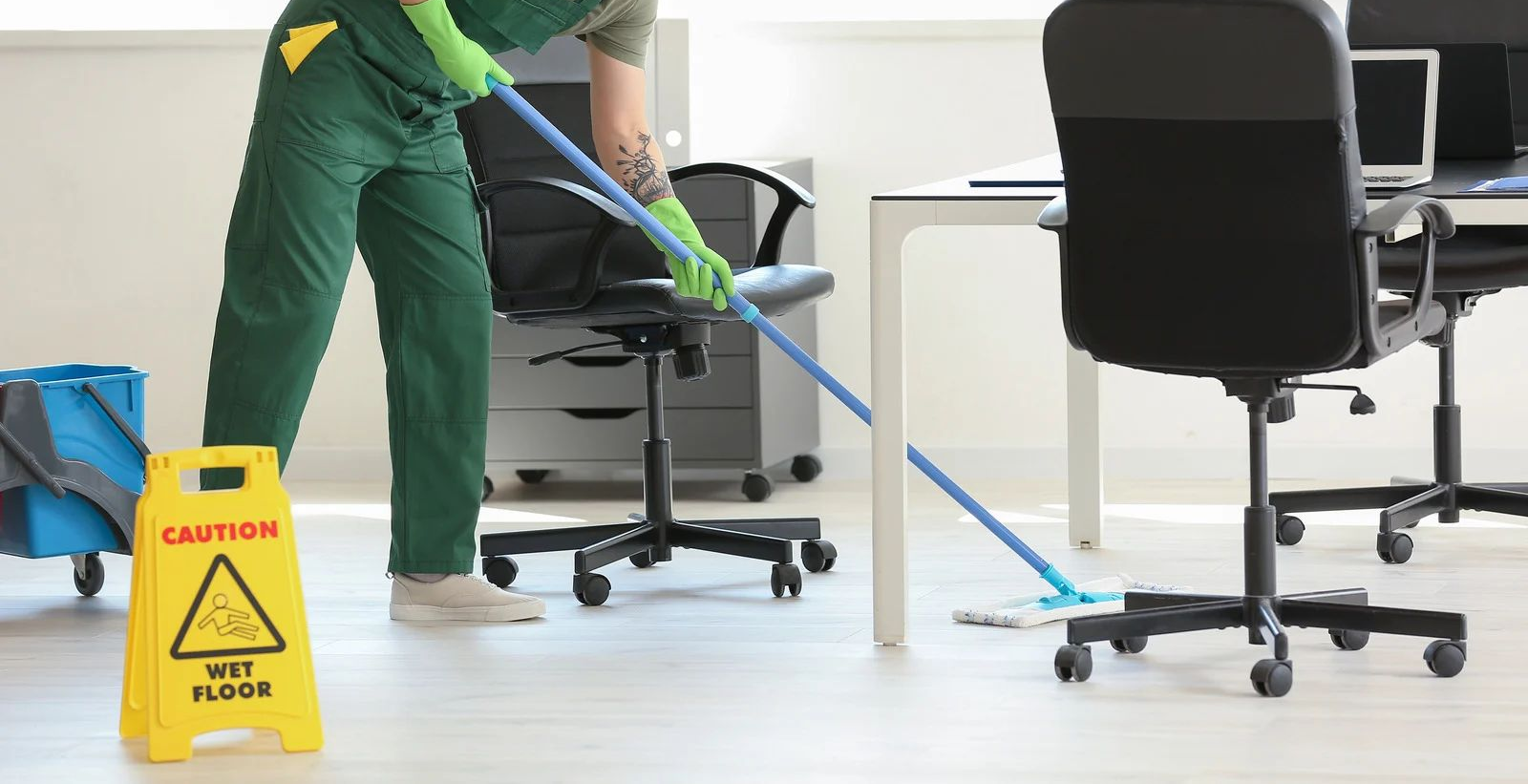 A person in green cleaning an office floor with a mop. A caution sign is in the foreground.