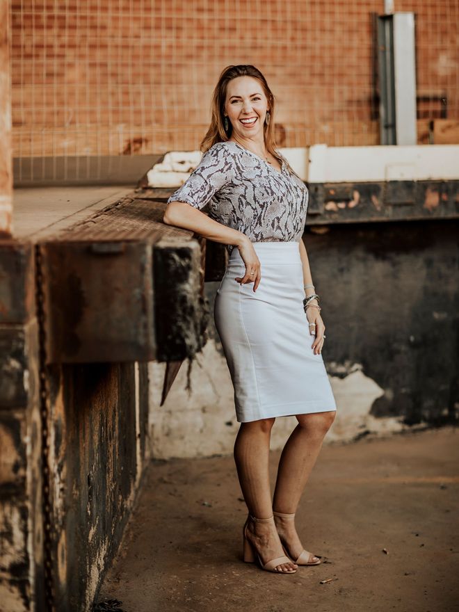 Woman in a floral blouse and white skirt leaning on a concrete wall in an industrial setting.