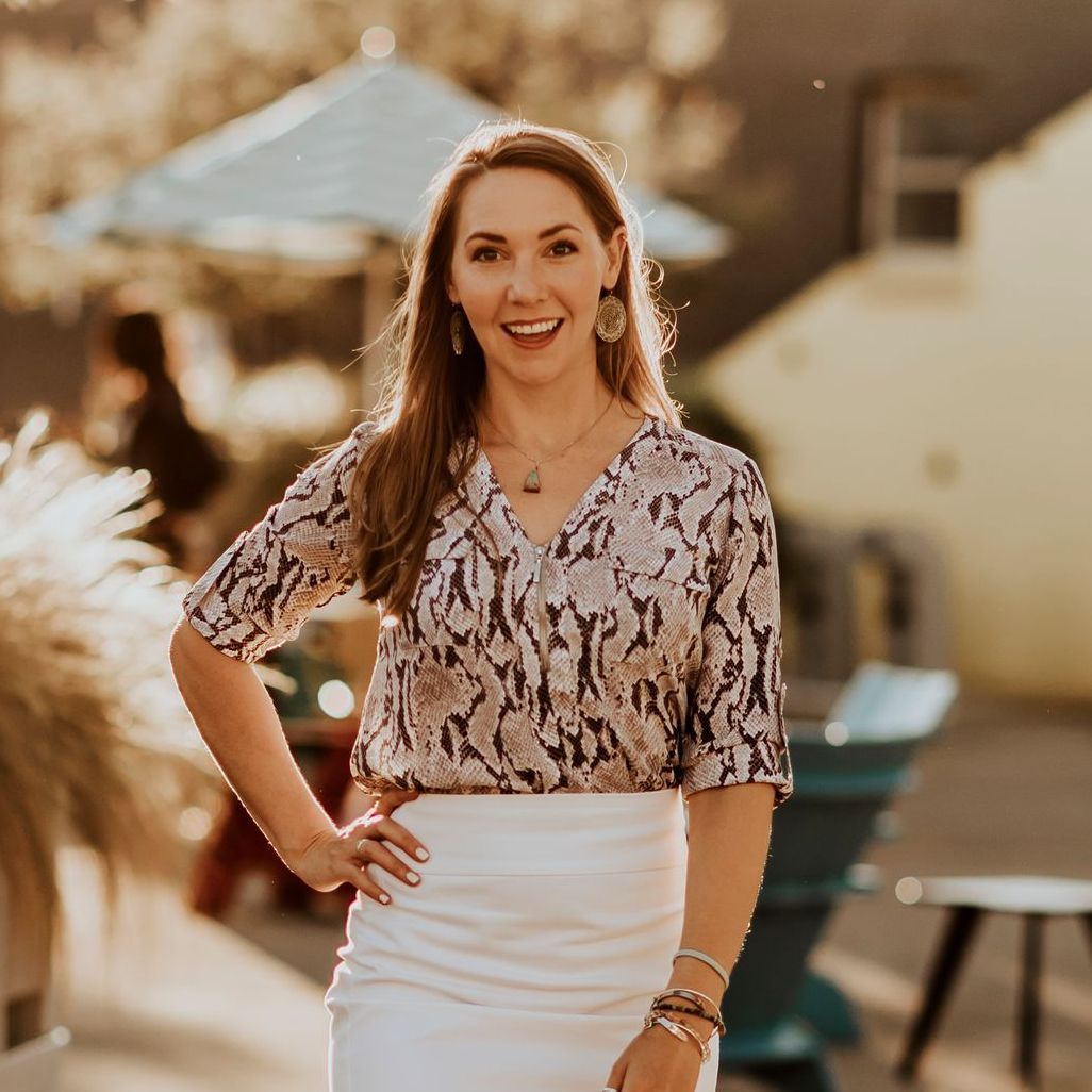 Woman smiling outdoors in a patterned blouse and white skirt, standing in a sunny patio setting