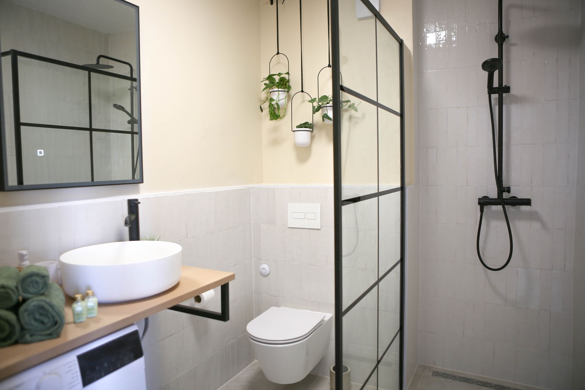 Modern bathroom with black framed shower, vanity, and hanging plants.