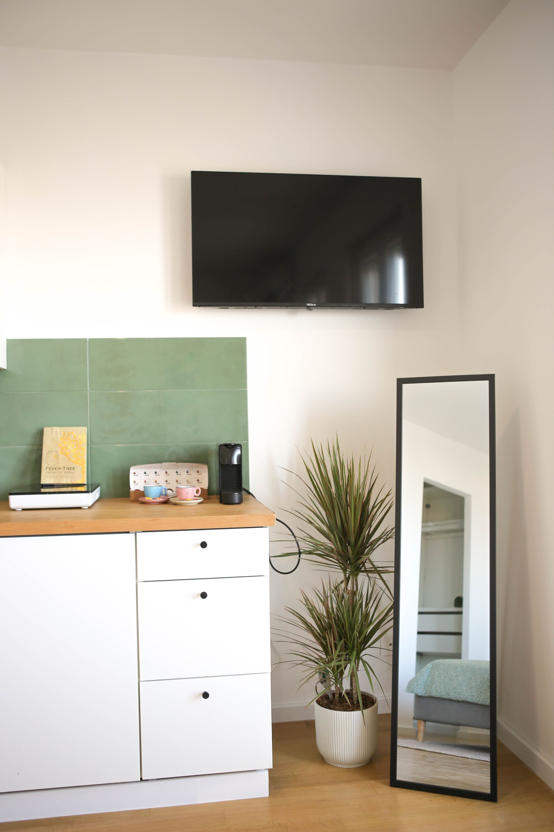 White cabinet with tv, mirror, and plant. Green tile backsplash, light wood floor, neutral walls.