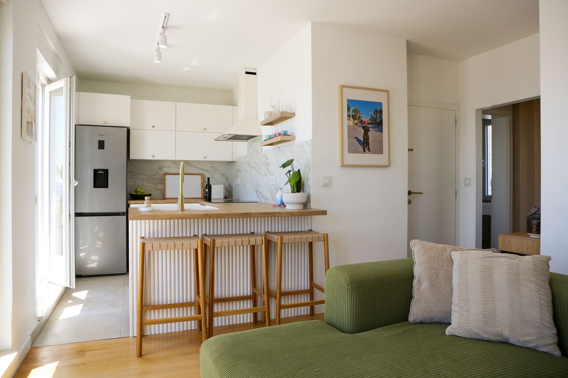 Bright, modern kitchen and living space. Green sofa, wood bar stools, white cabinets, and stainless steel fridge.