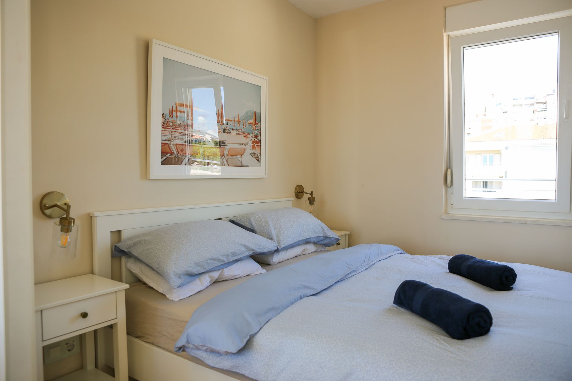 Bedroom with white bed, blue bedding, artwork, window, and nightstand.