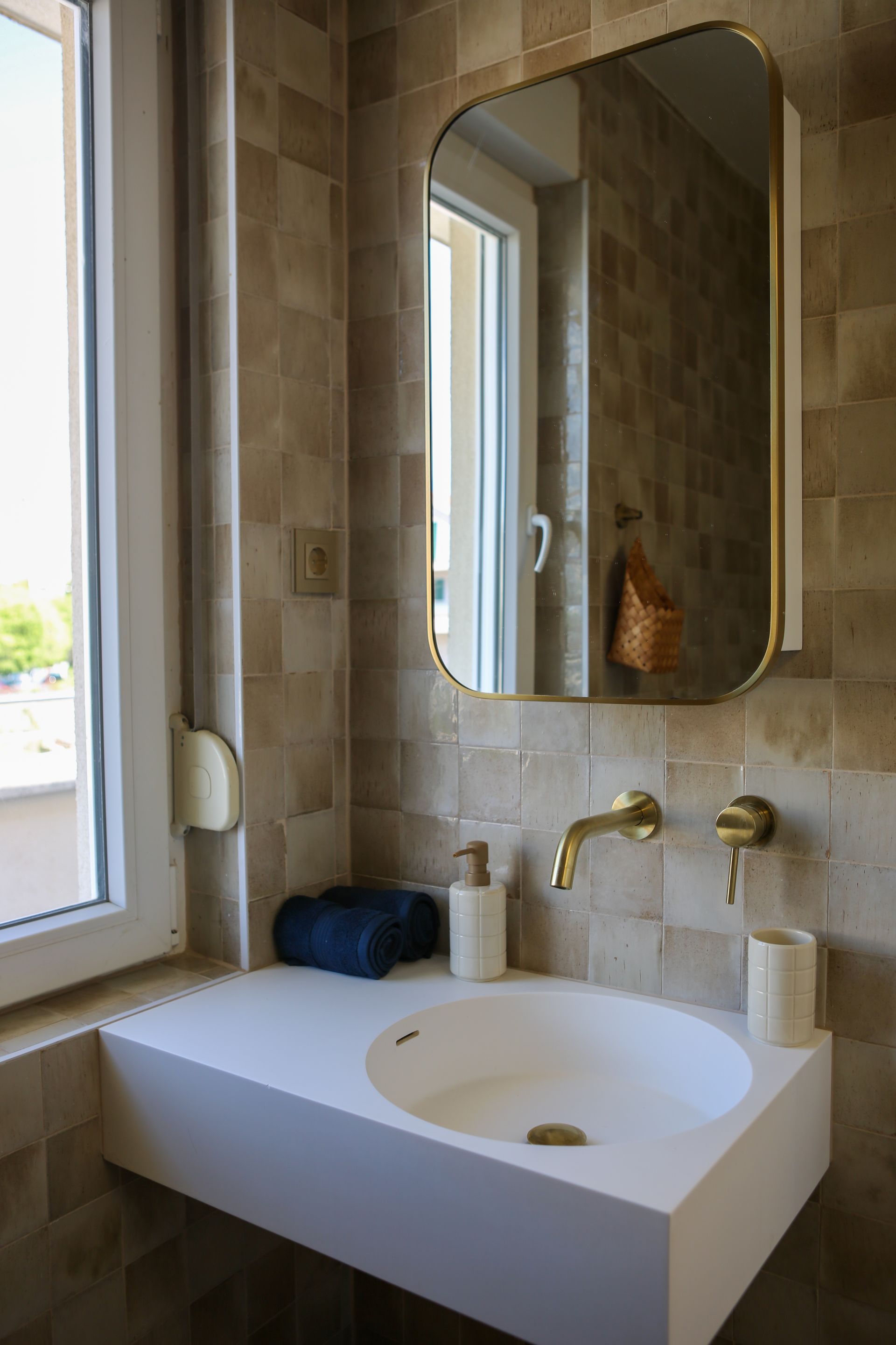 Bathroom with square tile walls, a white sink, gold faucet and mirror, and a window.
