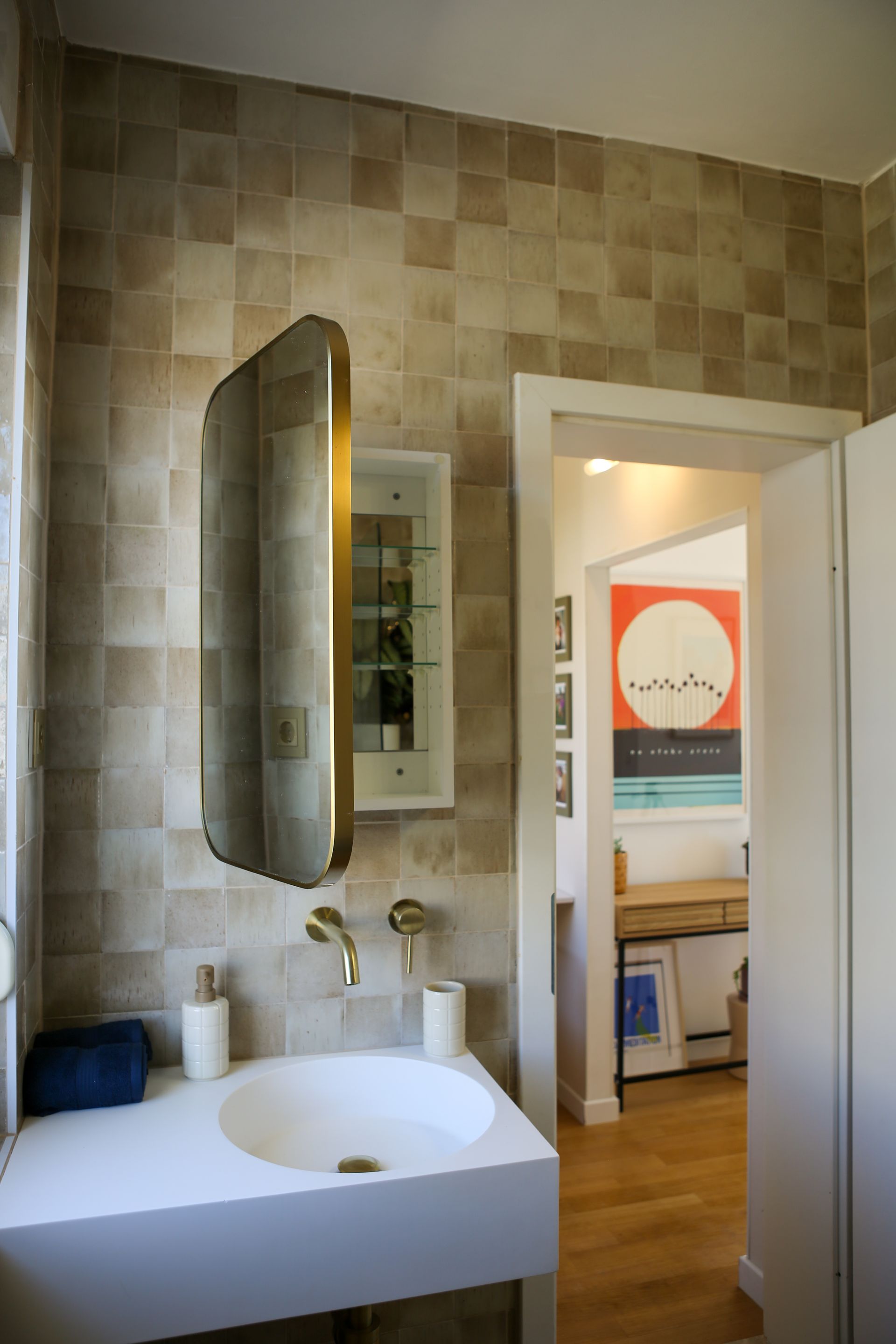 Bathroom with white sink, gold faucet and mirror, and tan square tile walls. Doorway to another room.