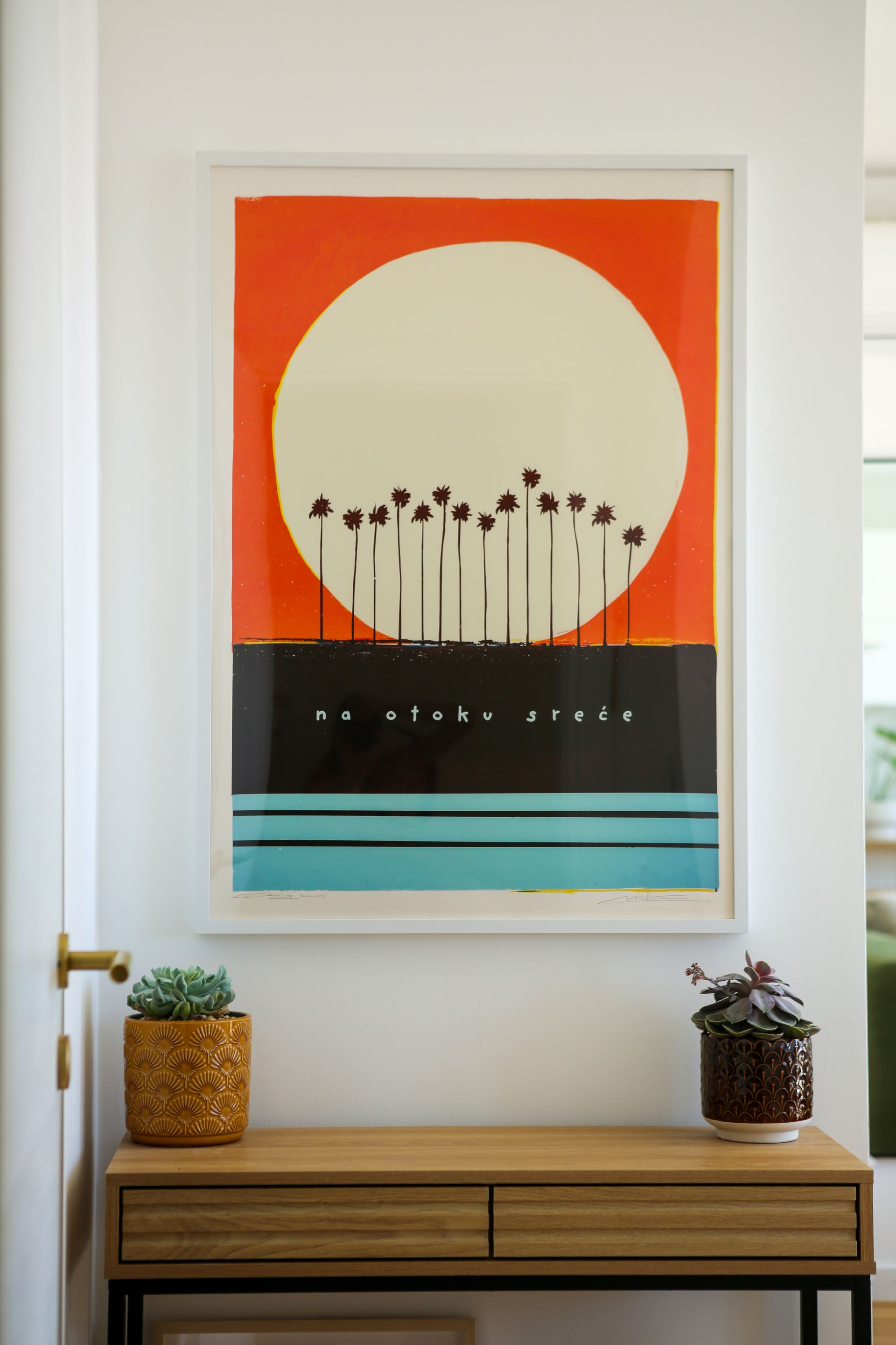 Artwork of a sunset over silhouetted palm trees, above a blue line, on a white wall.