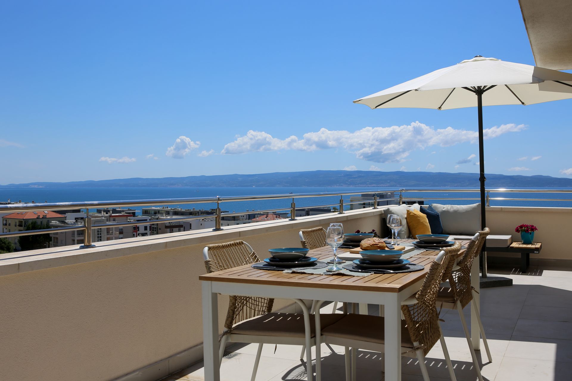 Rooftop dining table set for a meal, overlooking city and sea under a sunny blue sky.