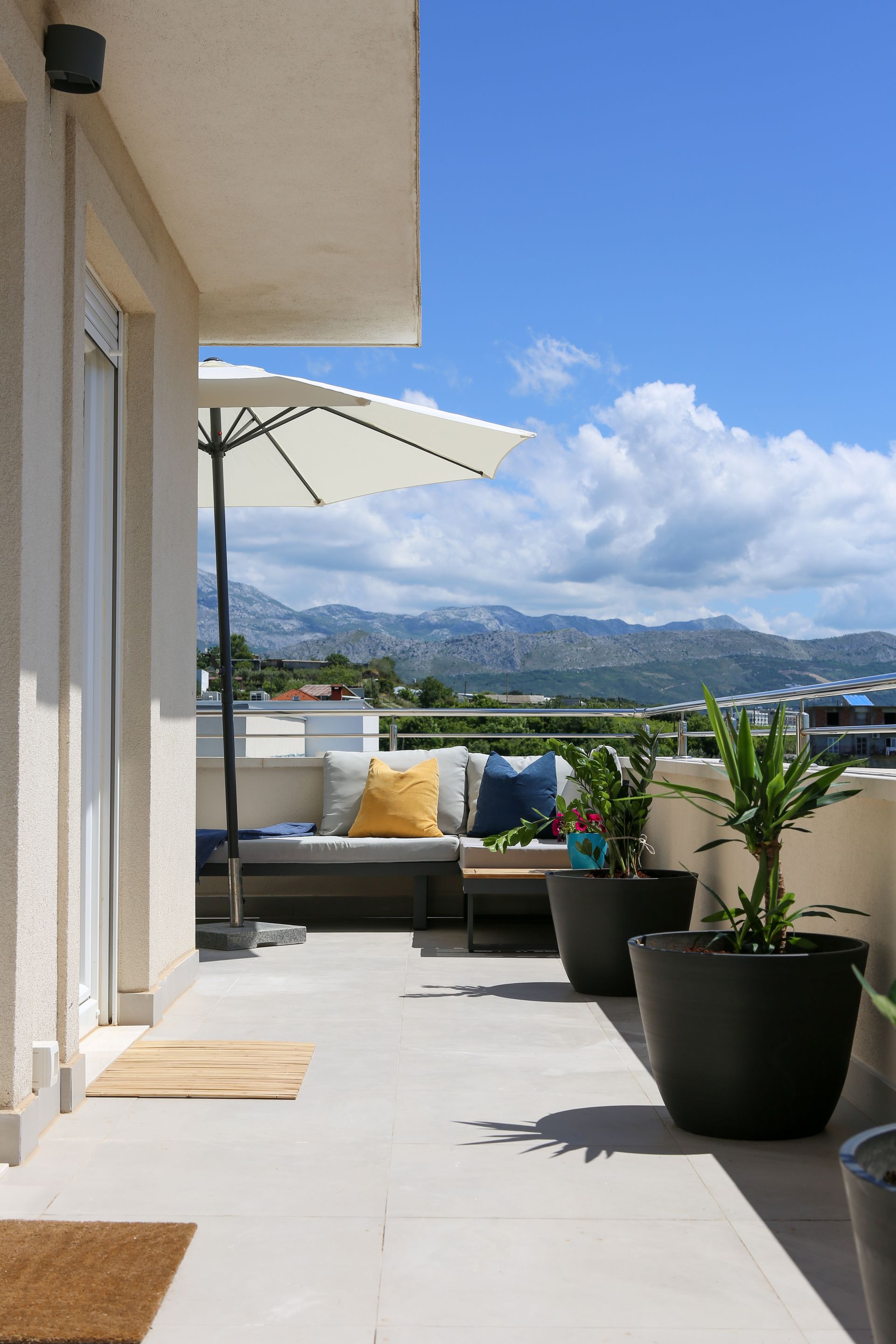 Balcony with potted plants, cushioned seating, umbrella, and mountain view under blue sky.