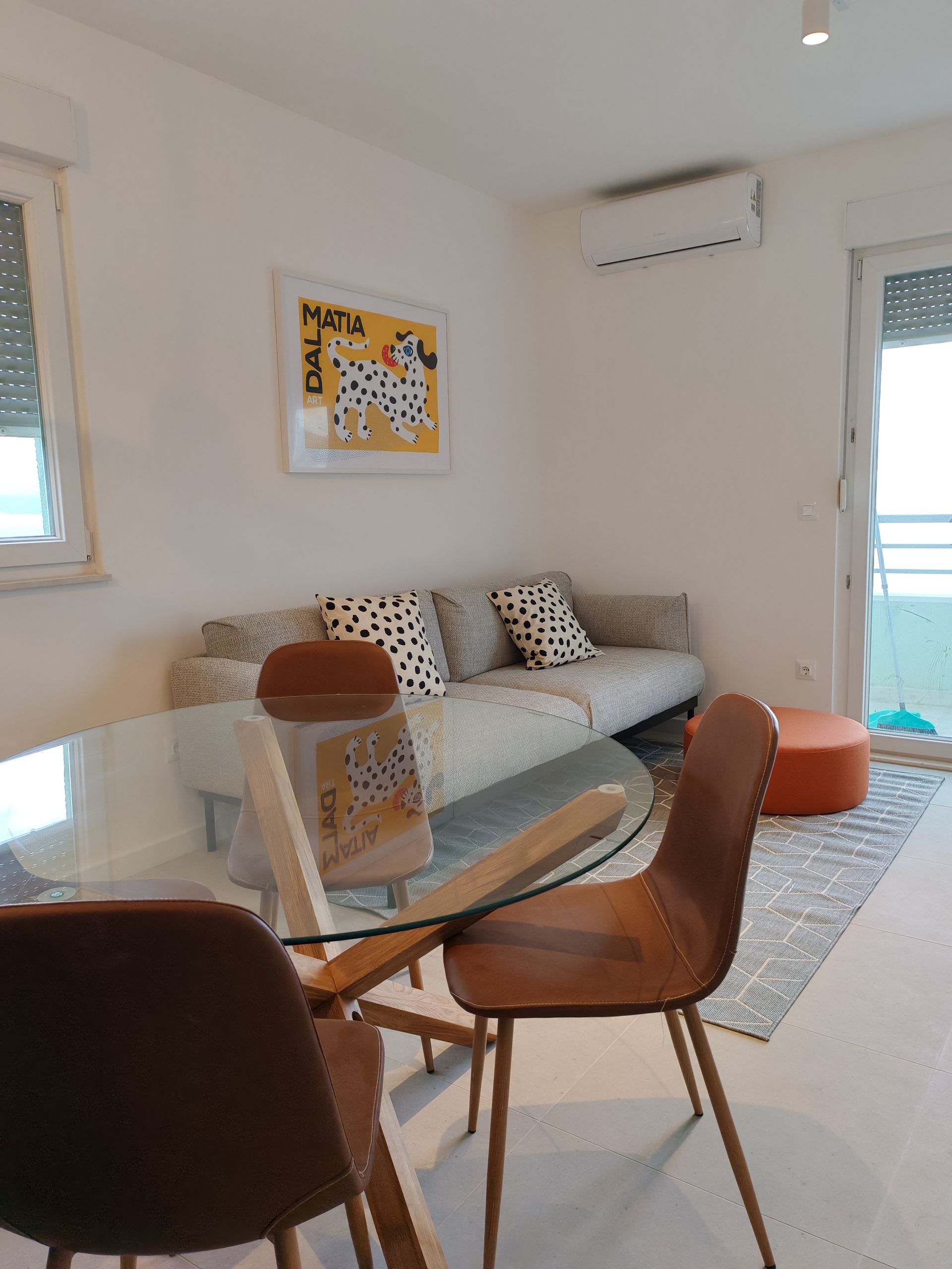 Living room: sofa, round glass table with chairs, Dalmatian art, air conditioner, and orange ottoman.