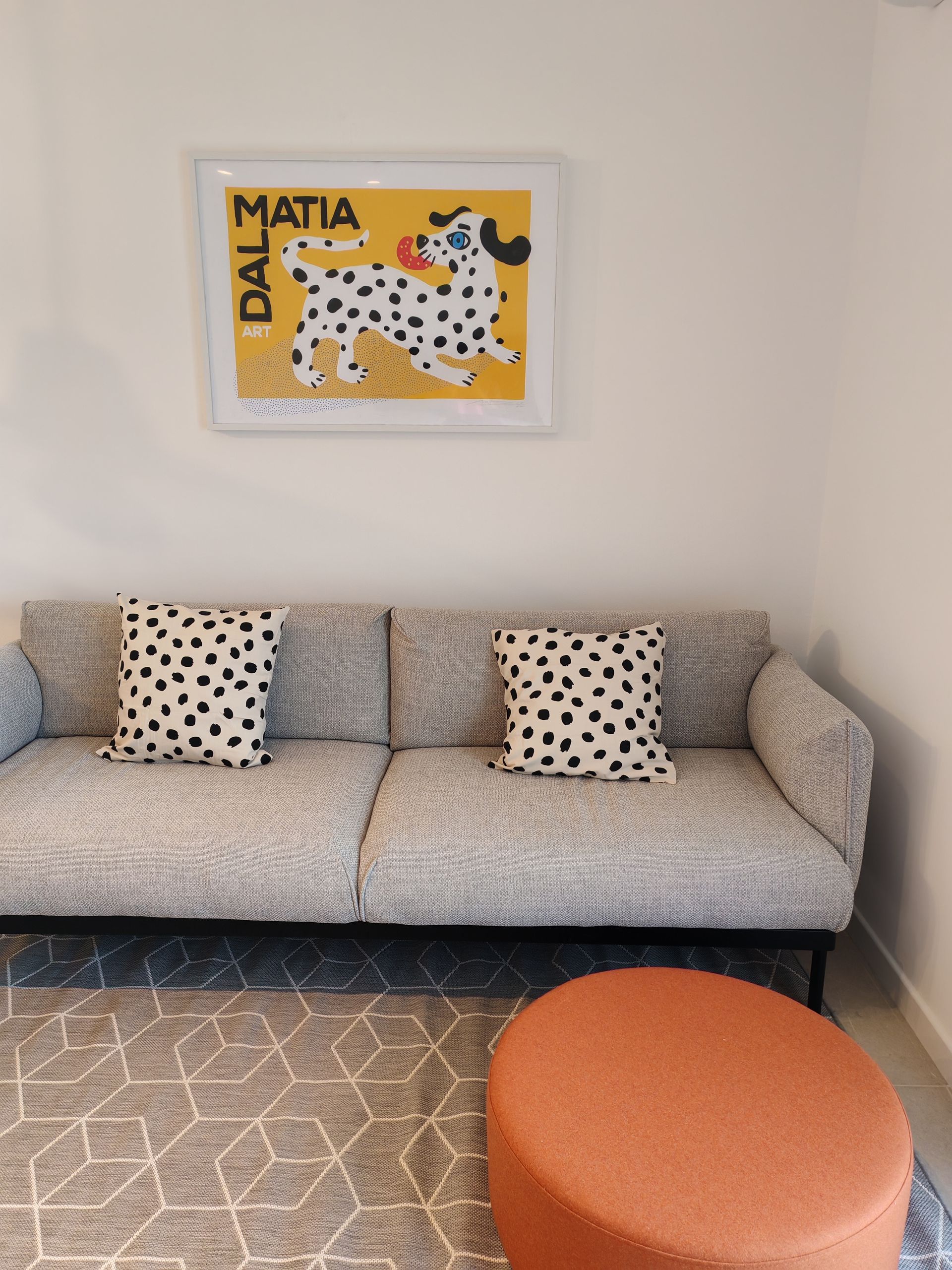 Couch with dalmatian-print pillows, orange ottoman, and a framed dog art piece on a white wall.