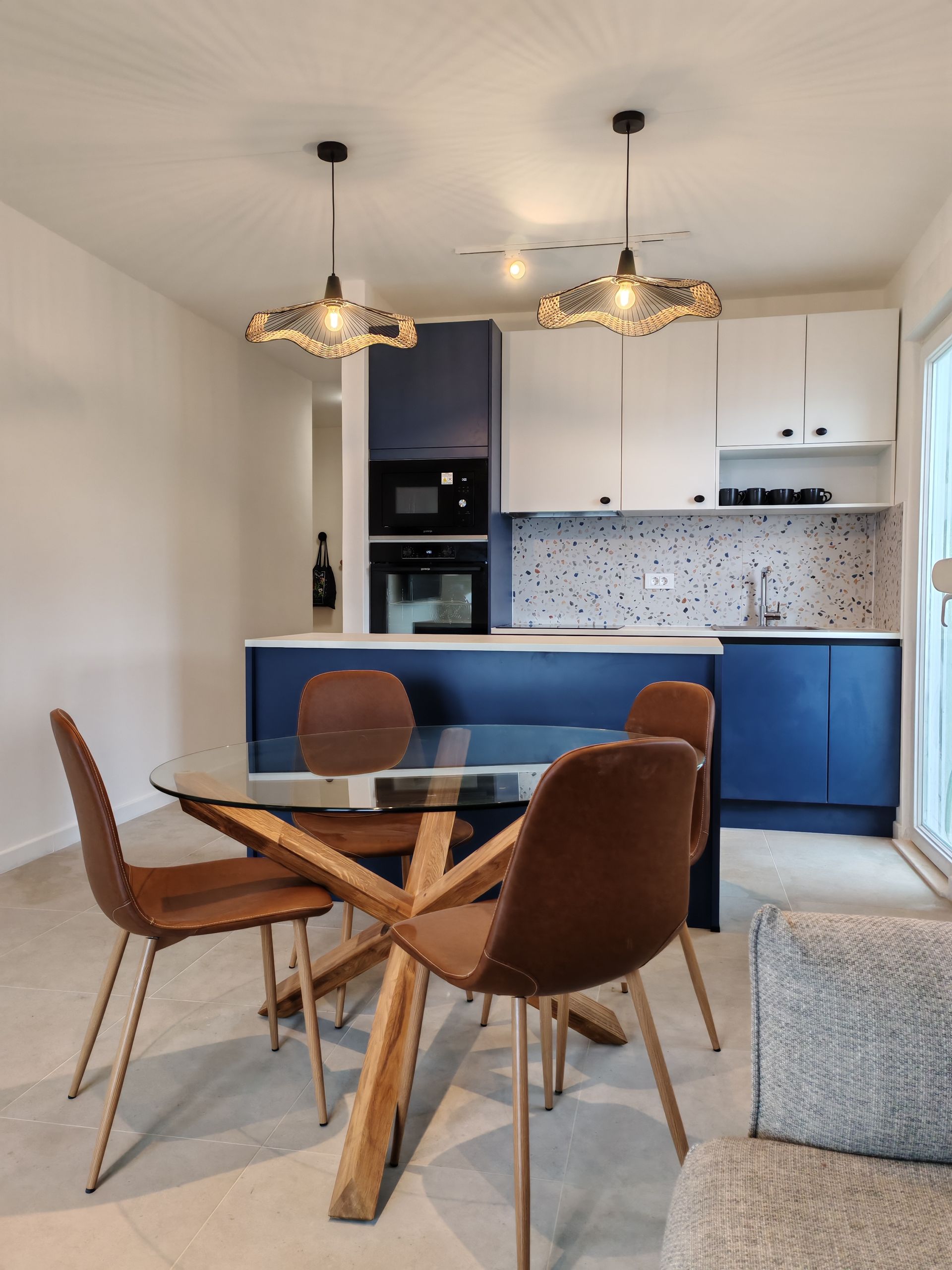 Modern kitchen and dining area with blue and white cabinets, glass table, brown chairs, and decorative lights.