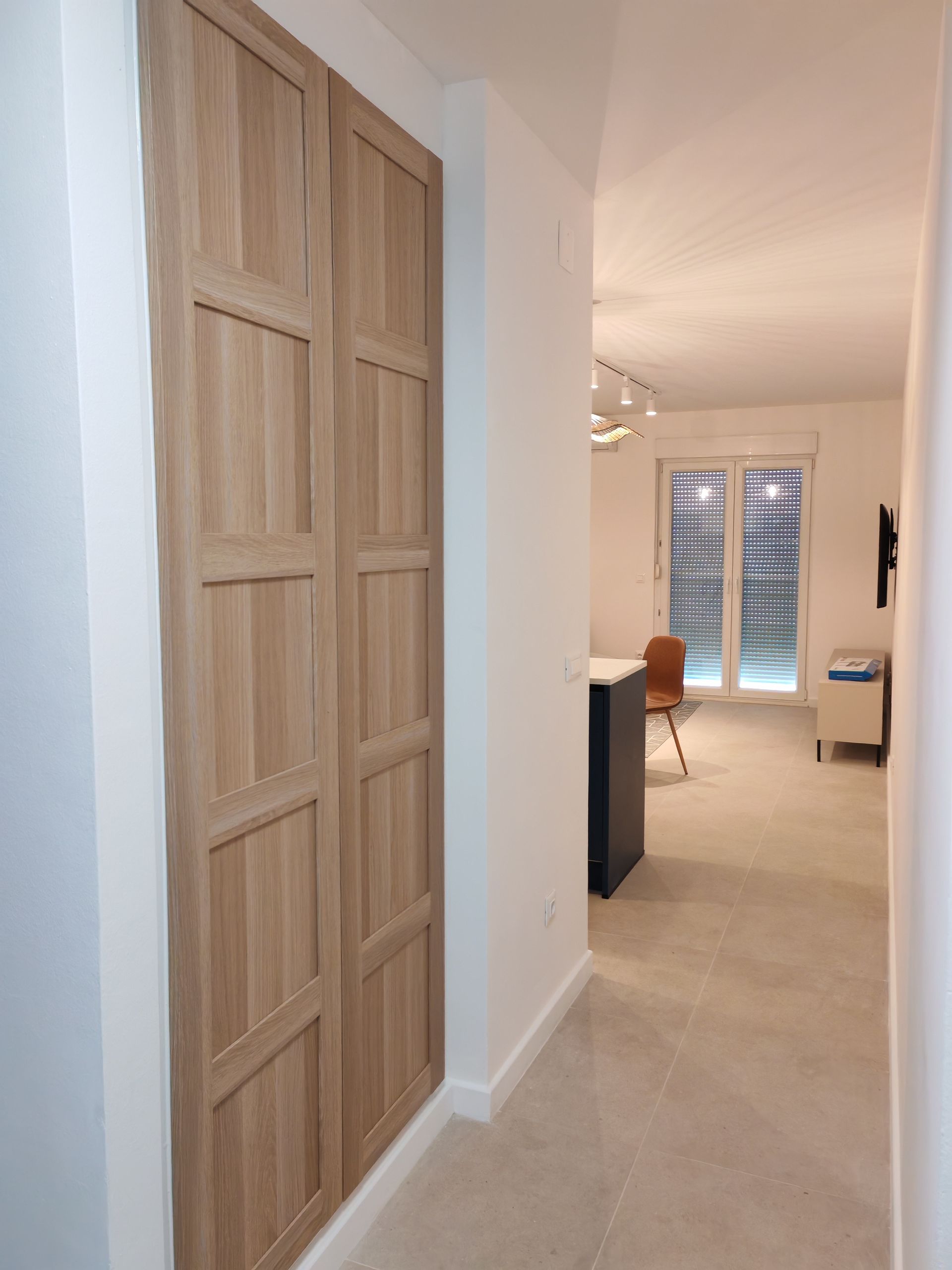 Wooden closet doors in a narrow hallway, leading to a living area with a window and a bar.