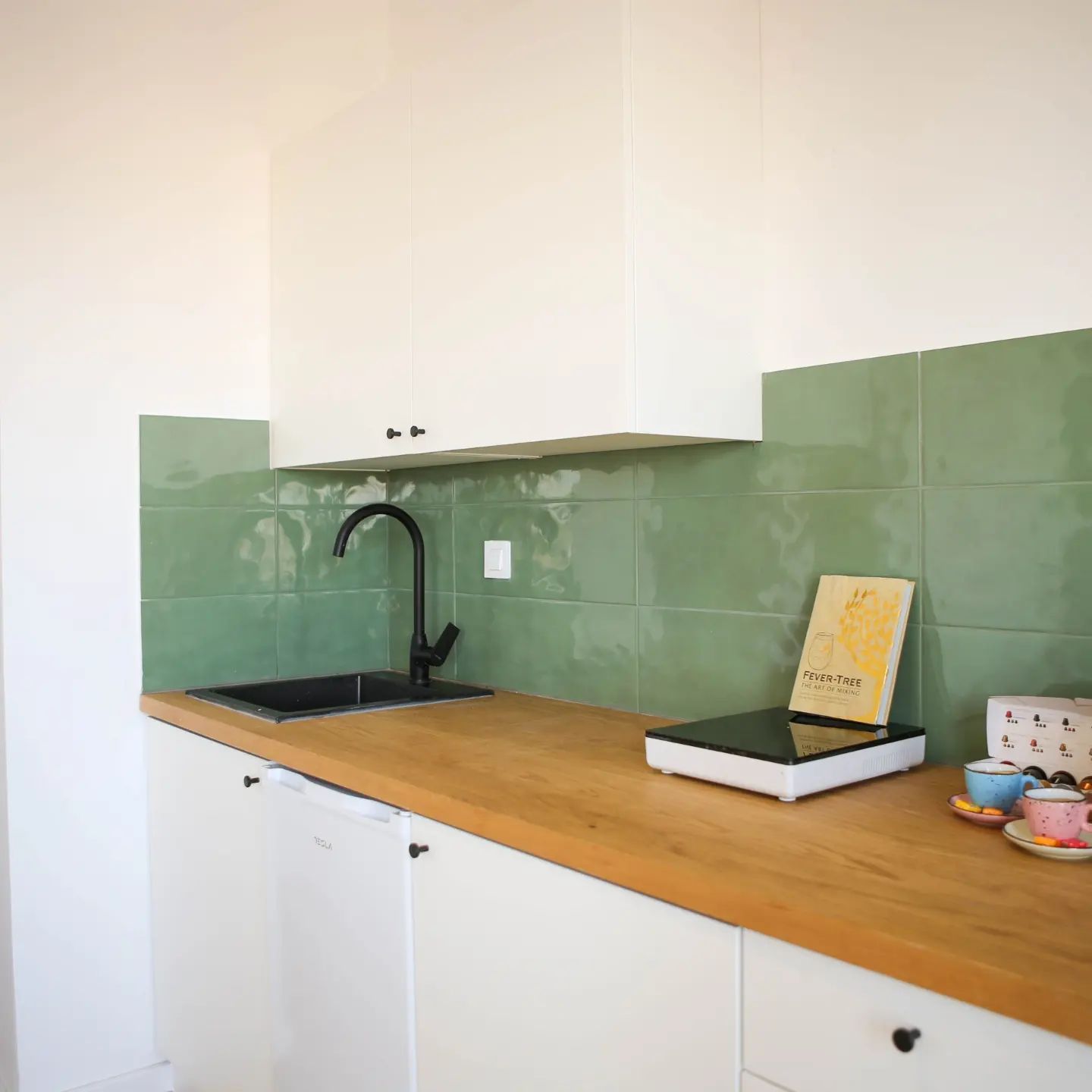 White kitchen with wooden countertop, black sink and faucet, green backsplash.