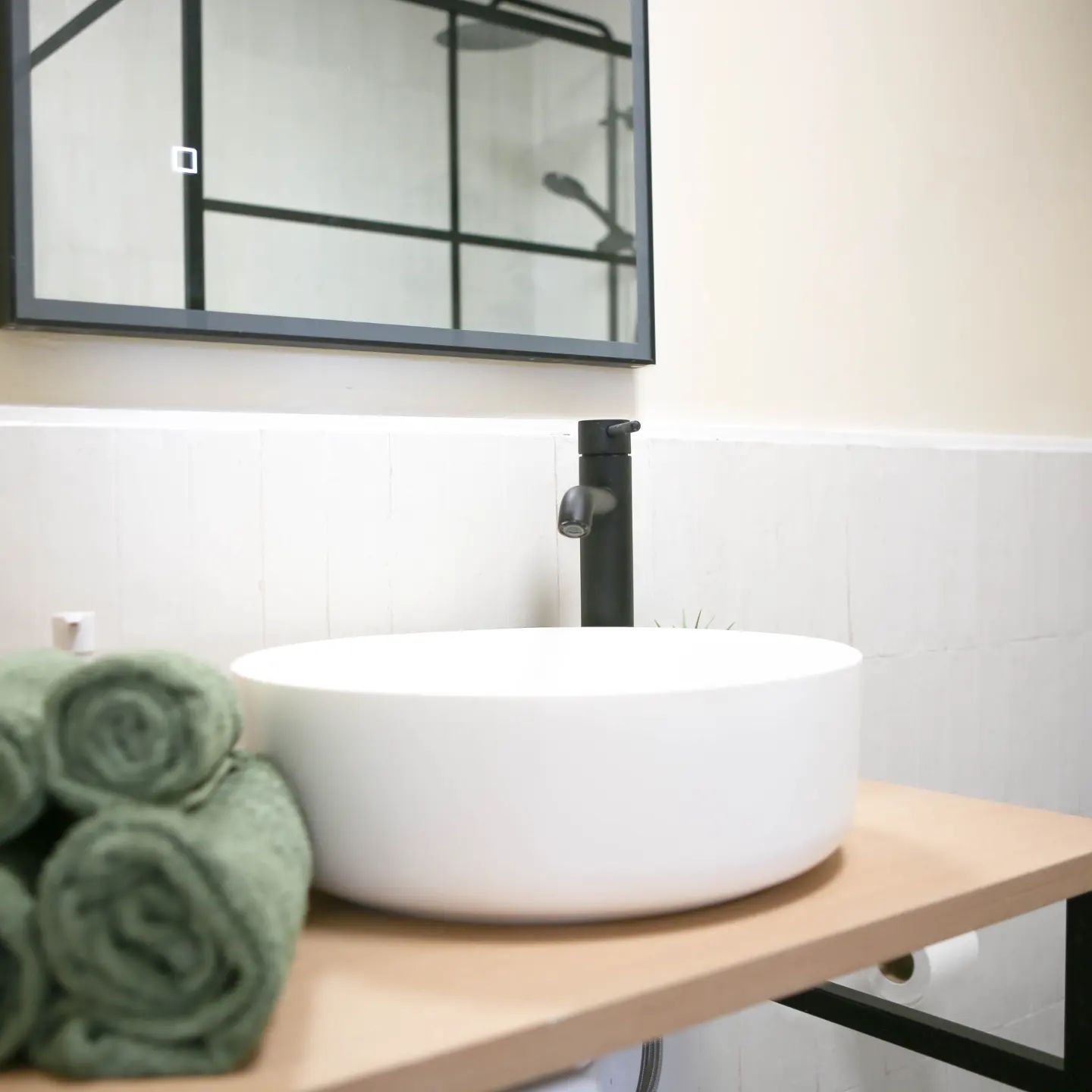 White bowl sink on a wooden counter with green towels, black faucet, and a large framed mirror.
