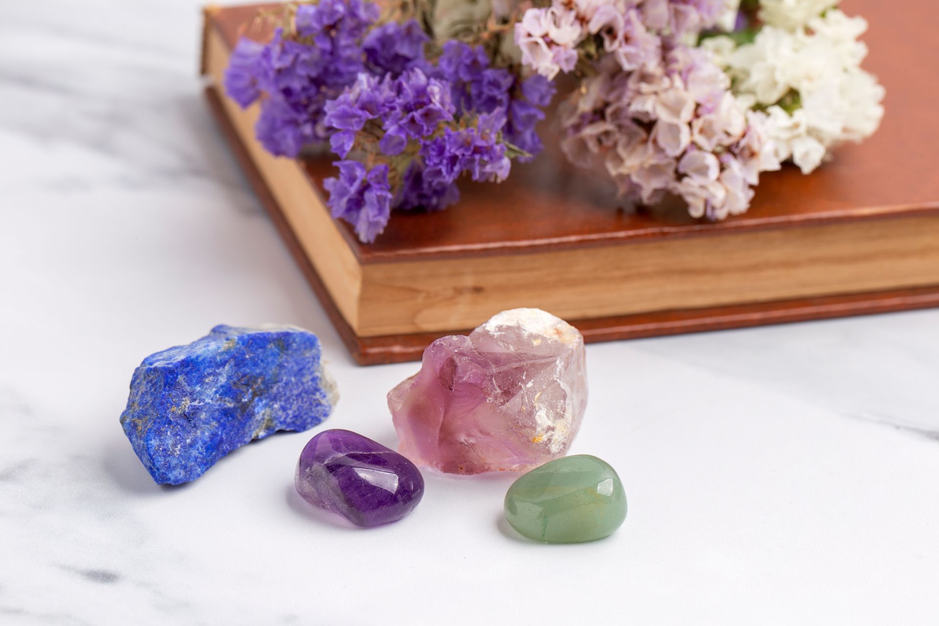 Des cristaux bleus, violets et verts à côté d'un livre et de fleurs séchées sur une surface en marbre blanc.