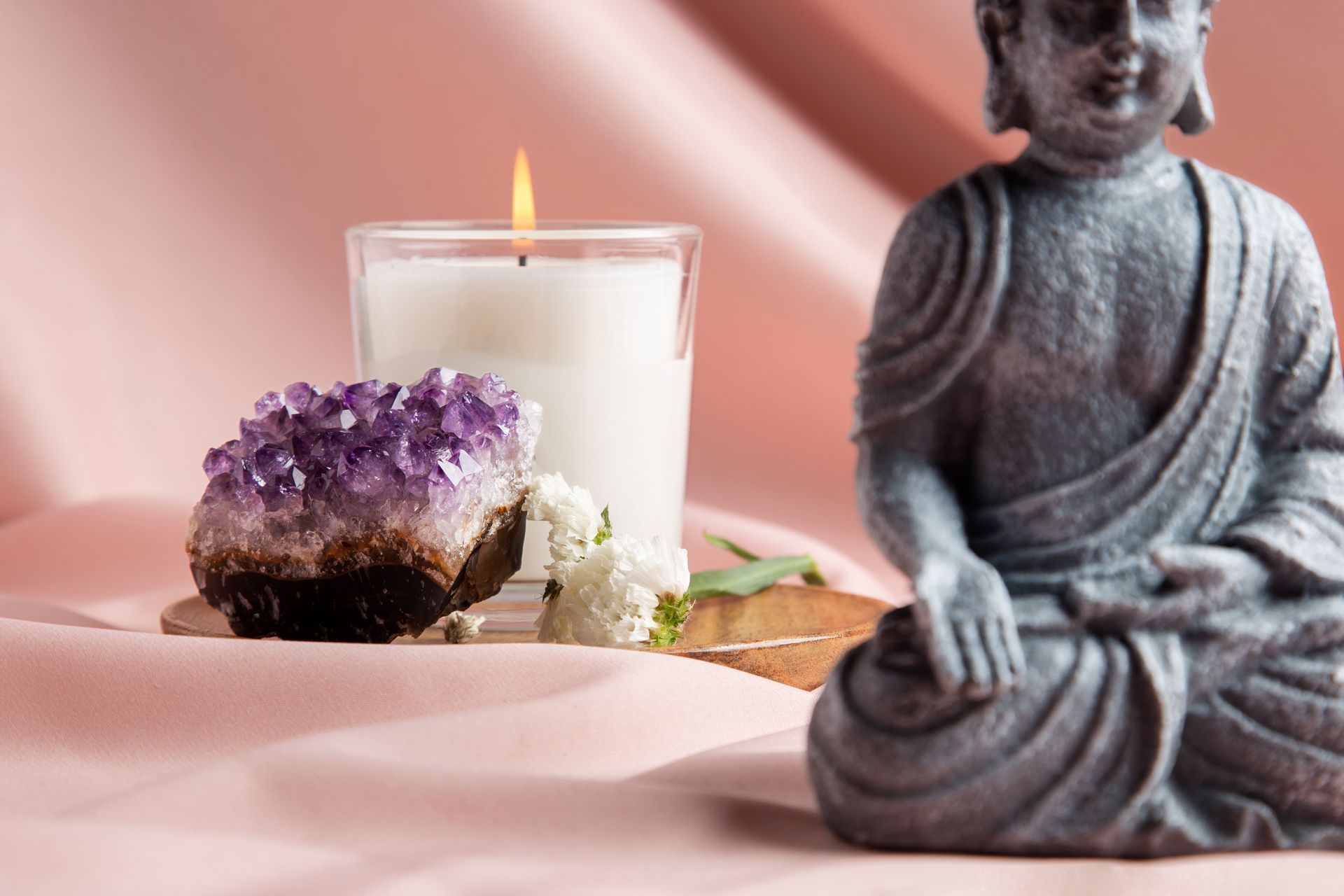 Statue de Bouddha, bougie allumée, amas d'améthystes et fleurs sur tissu rose.