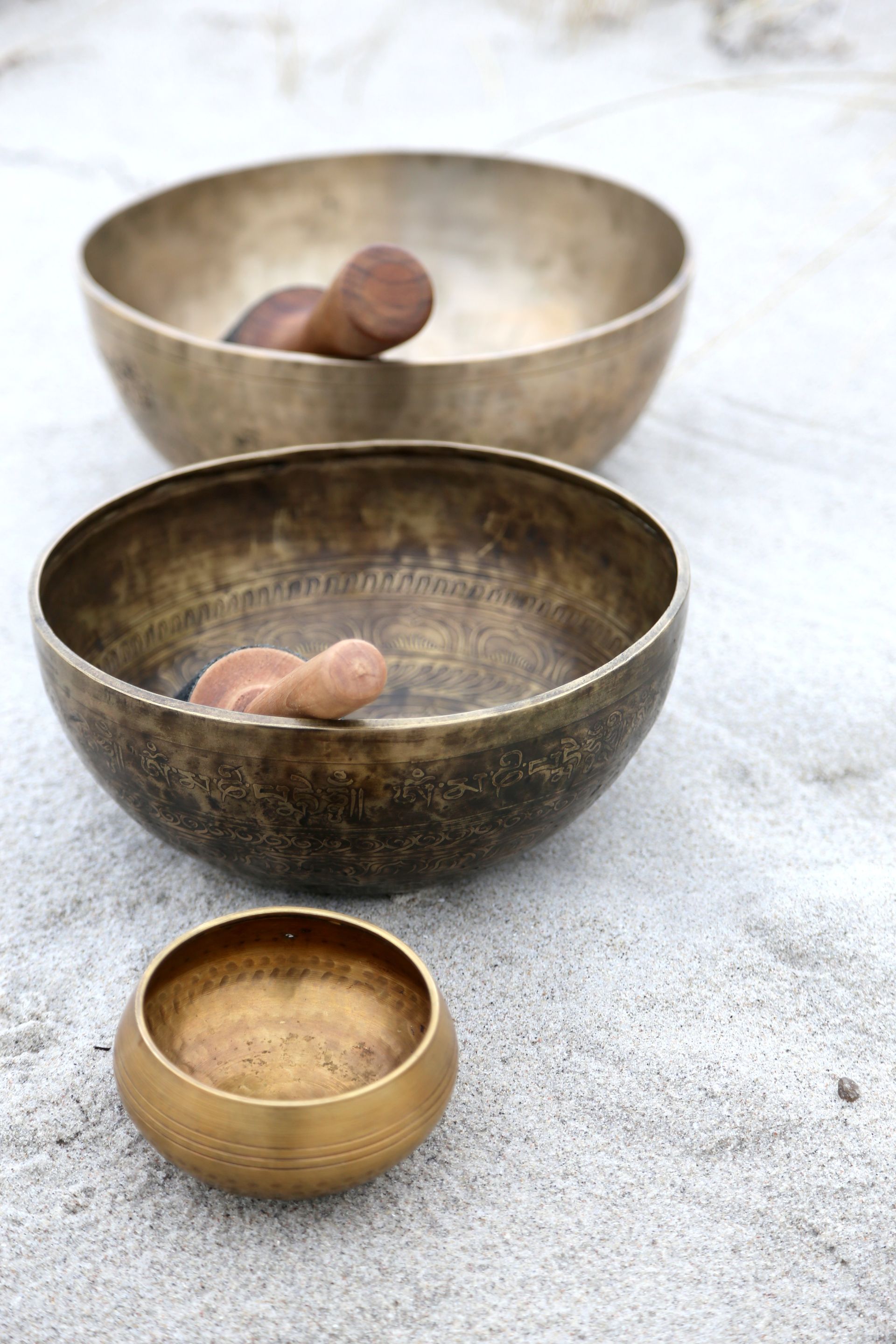 Trois bols chantants en bronze avec des maillets en bois, sur une surface sablonneuse.