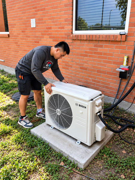 Technician inspecting an outdoor air-conditioning unit beside a brick house.