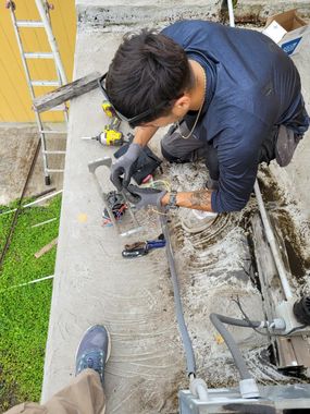 Worker repairing electrical wiring on a concrete ledge beside a yellow wall, with tools and a ladder nearby