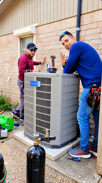 Two HVAC technicians working on an outdoor air conditioner unit beside a brick building.