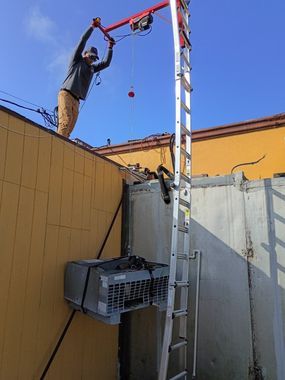 Worker on a ladder using a red rope hoist to lift equipment over a rooftop wall.