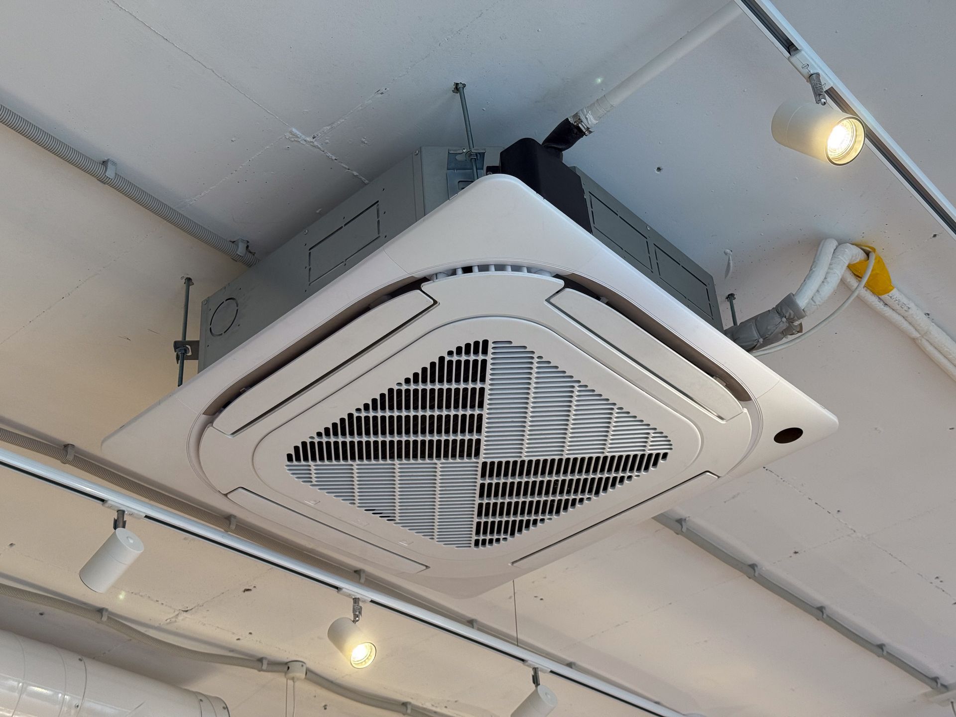 Ceiling-mounted cassette air conditioner in a white industrial ceiling with exposed pipes and lights