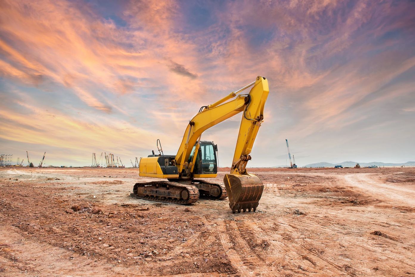 Escavatore giallo in un cantiere sterrato sotto un cielo dai colori vivaci al tramonto.