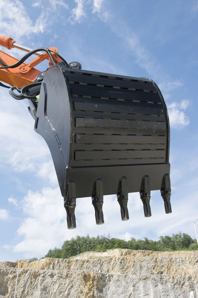 Benna di un escavatore sospesa sopra un cantiere sabbioso, sotto un cielo azzurro