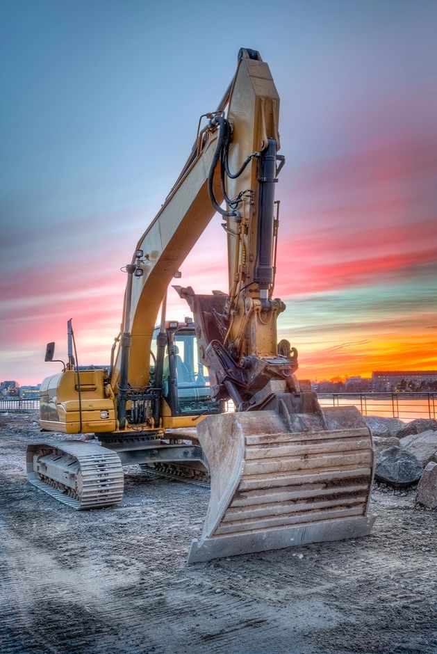Escavatore in un cantiere innevato al tramonto, con un cielo dai colori vivaci, rosa e arancione