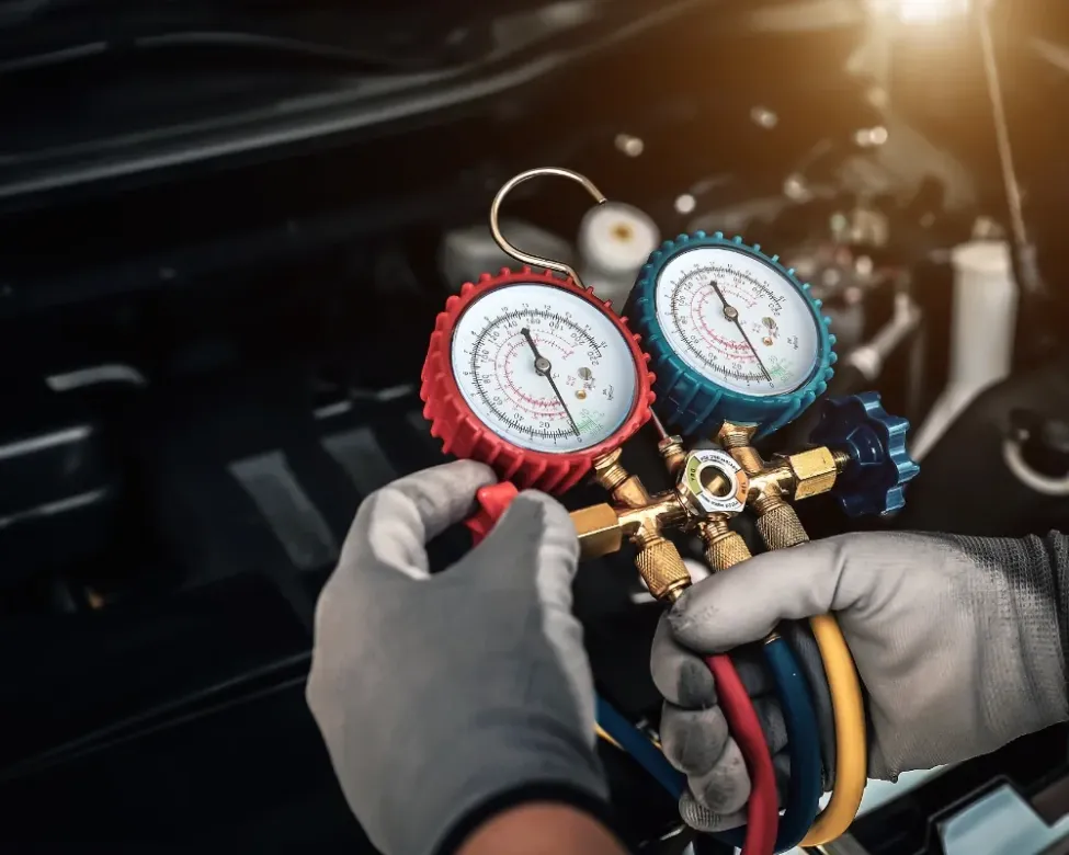 Mani guantate che tengono i manometri per l’aria condizionata dell’auto, con quadranti rossi e blu, sotto il cofano di un’auto