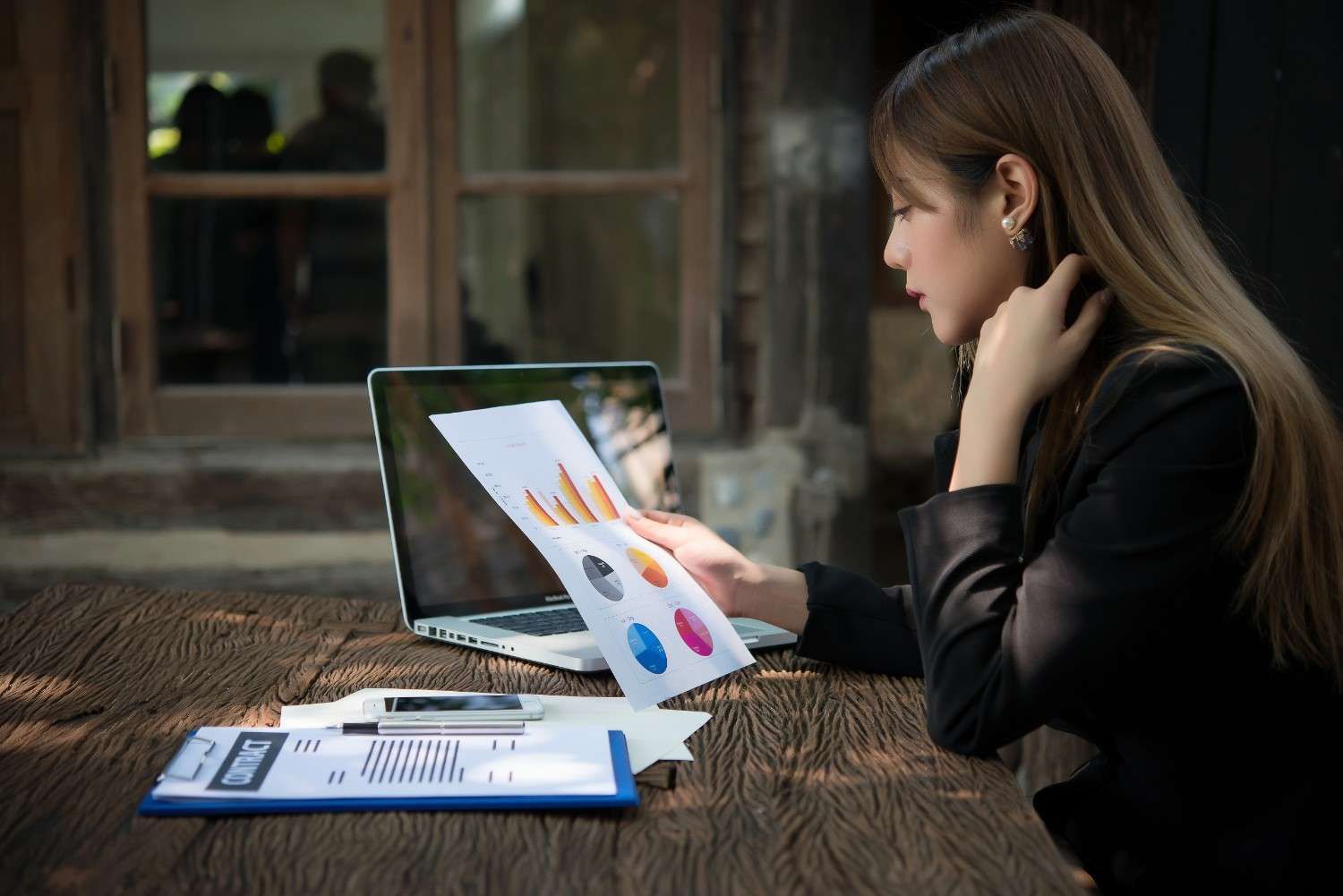 Person reviewing charts on a laptop at a wooden table, with papers and a tablet in a dim room