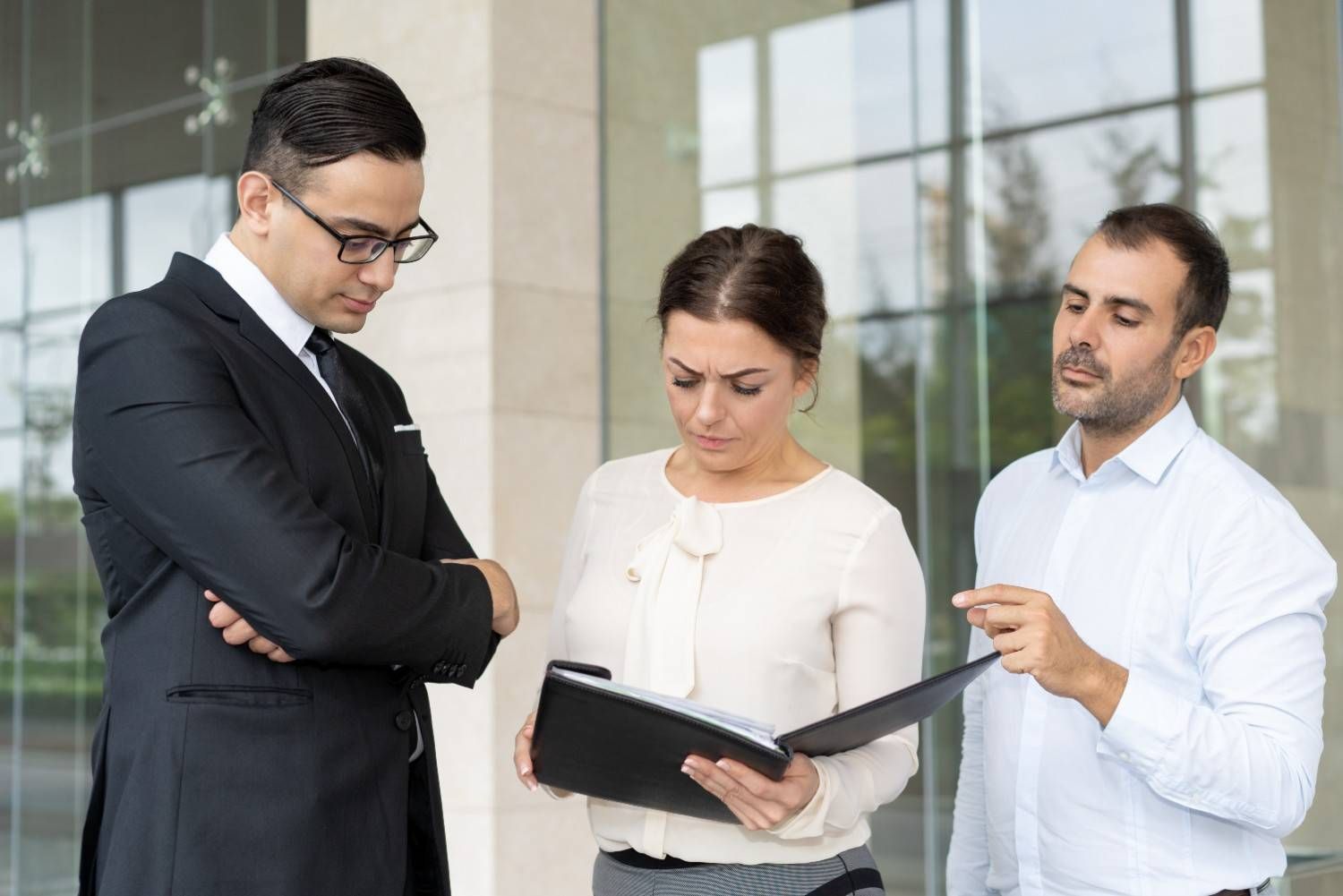 Three coworkers review a folder in a bright office lobby, one pointing while another takes notes.