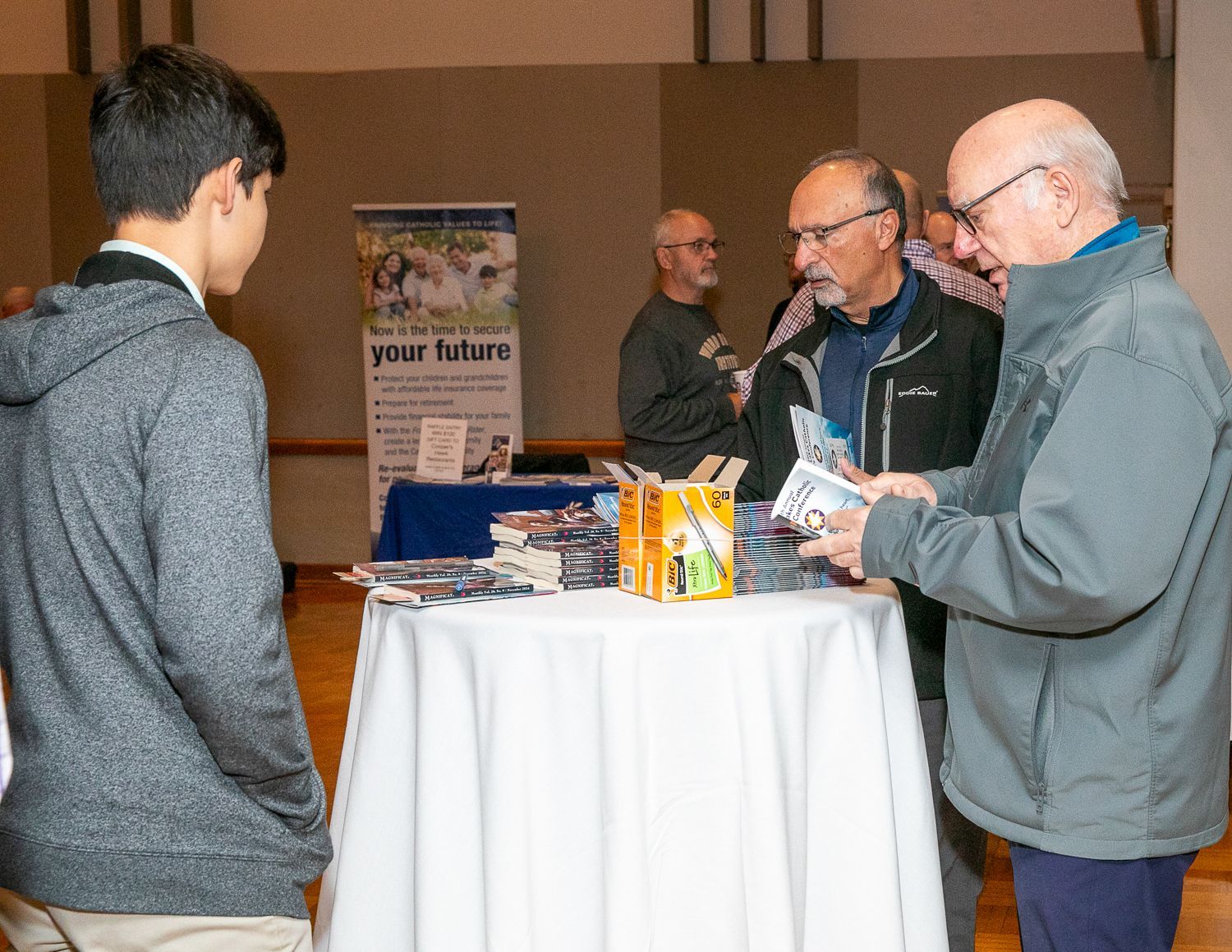 People talking at a small information booth with brochures and a sign in a lobby-like space