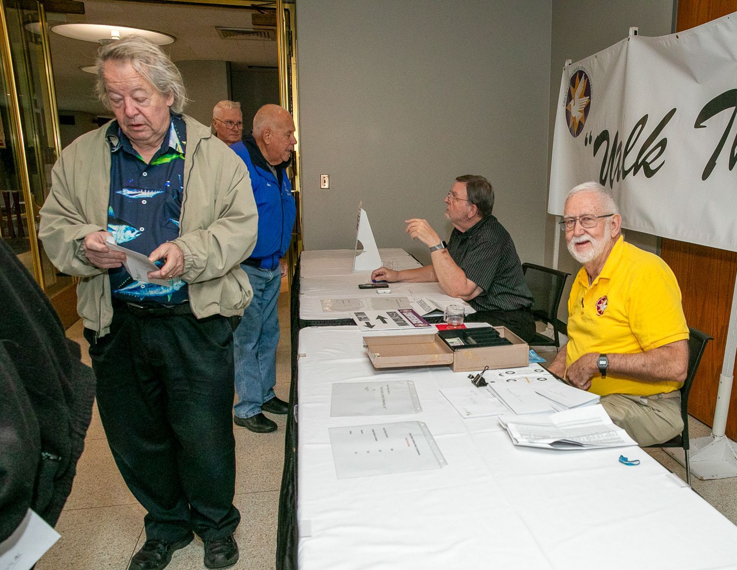 People at a registration table in a hallway, with paperwork and a man in a yellow shirt seated at the end.